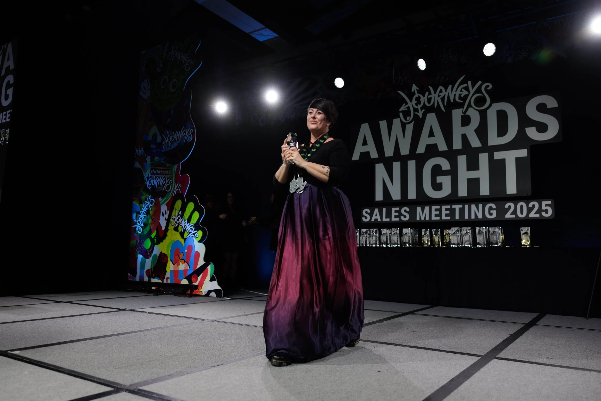 Woman standing on stage at Journeys Awards Night Sale Meeting 2025, holding an award, dressed in a black top and long purple skirt, with a large black and white sign behind her.