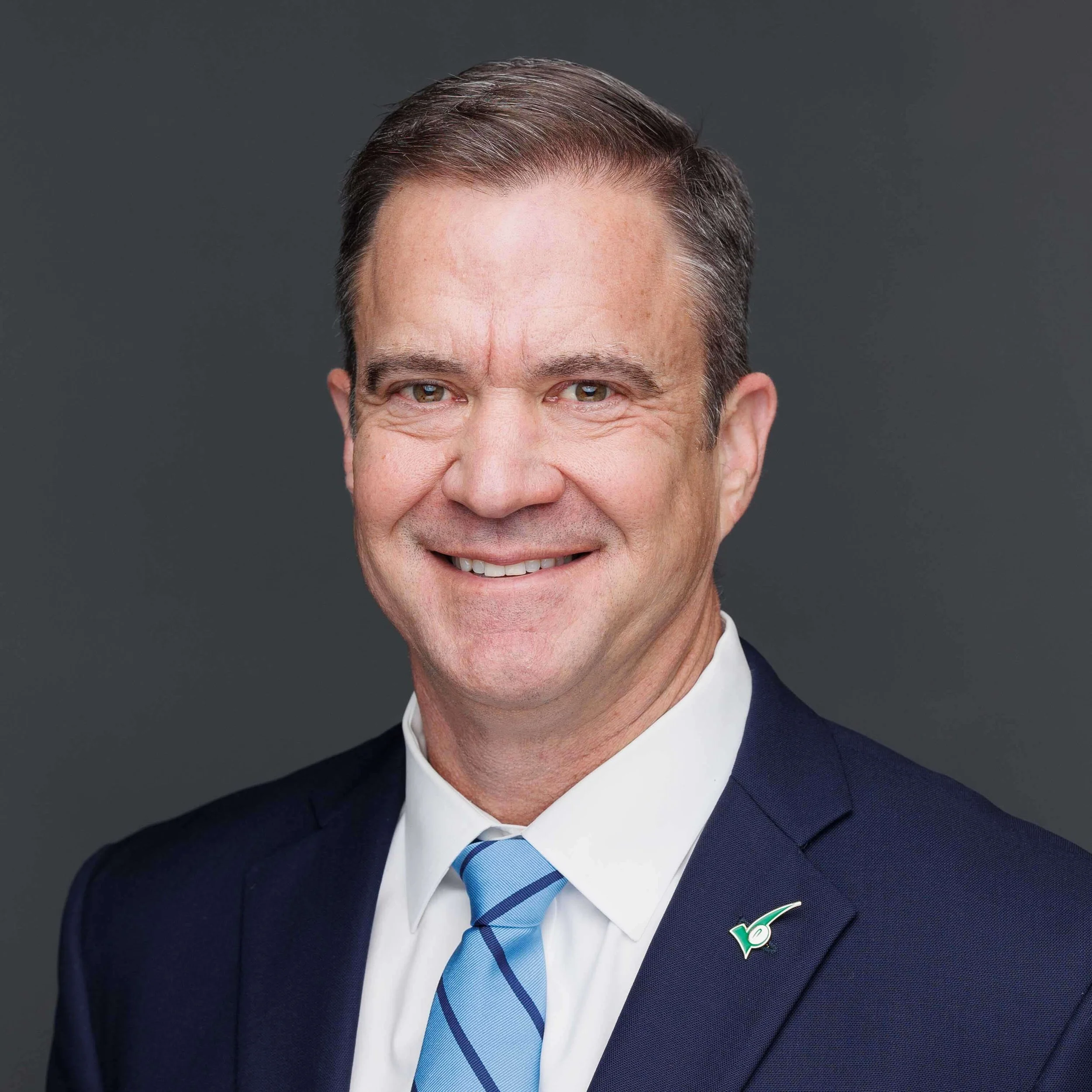A professional headshot of a middle-aged man in a dark suit with a blue striped tie, wearing a lapel pin, against a plain gray background.