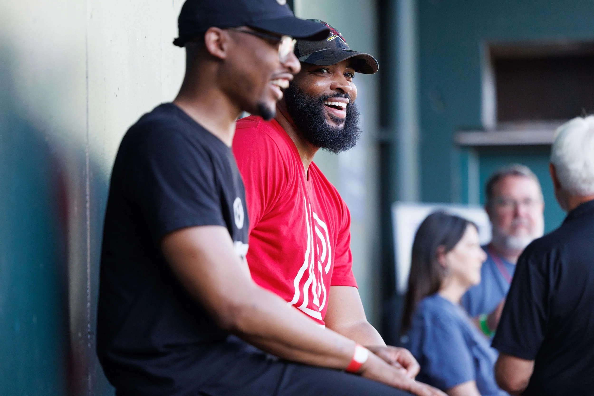 Two men sitting on a bench in a sports dugout, smiling and talking, with other people in the background at a sports event.