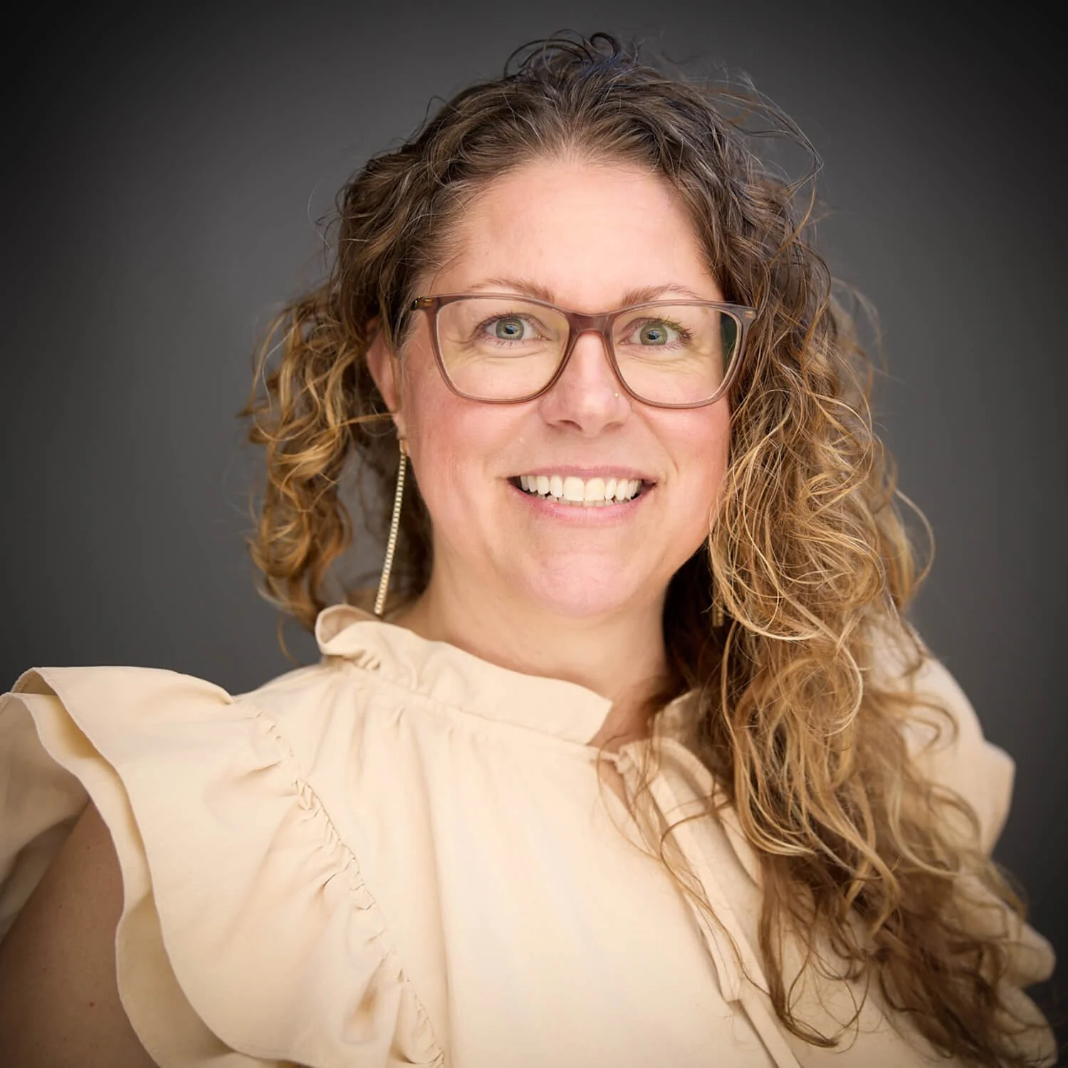 A woman with curly brown hair, glasses, and earrings smiling at the camera, wearing a cream-colored blouse with ruffled shoulders, against a dark gray background.
