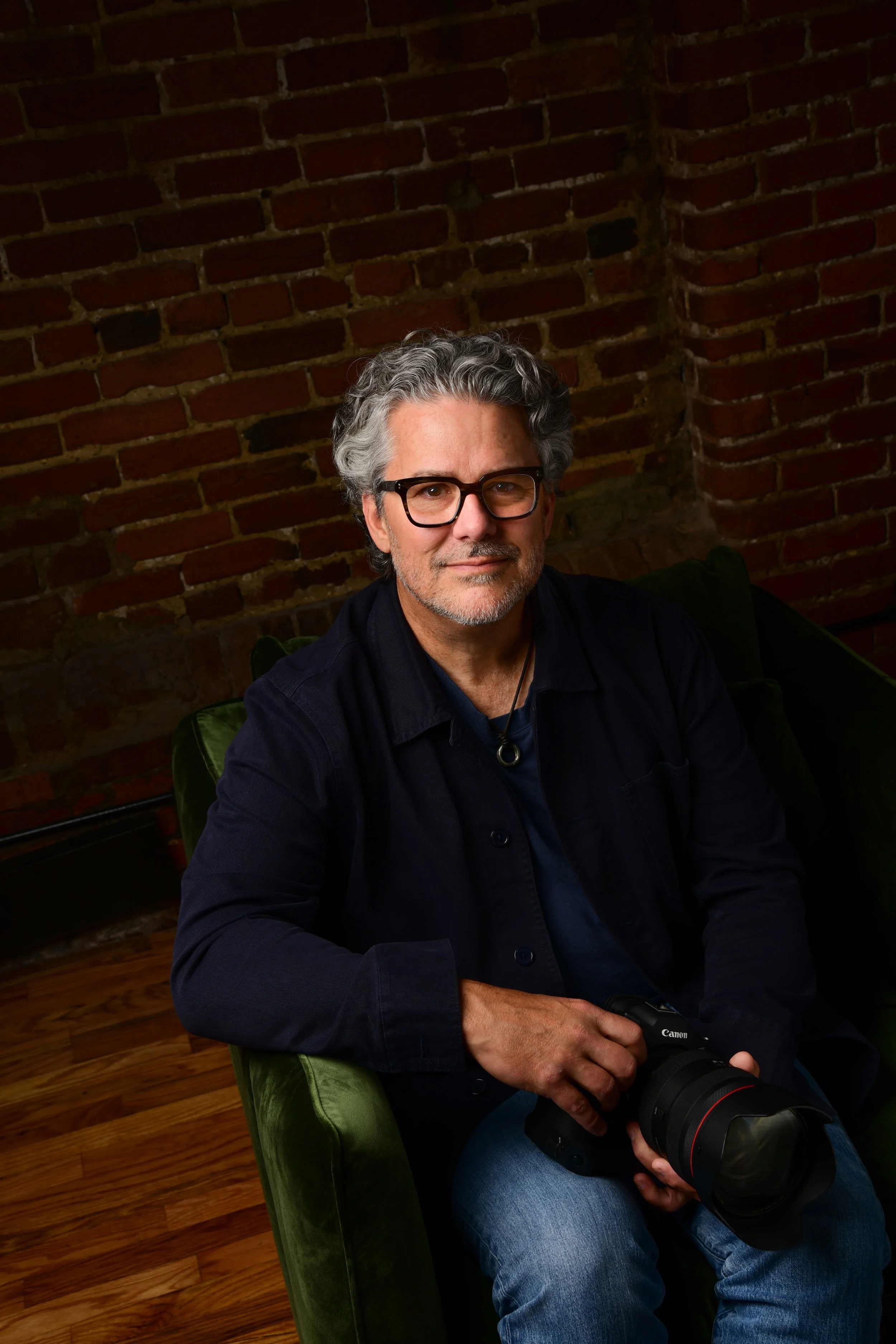 A middle-aged man with gray hair and glasses sitting on a green chair, holding a professional camera with a large lens, in a room with a brick wall background.