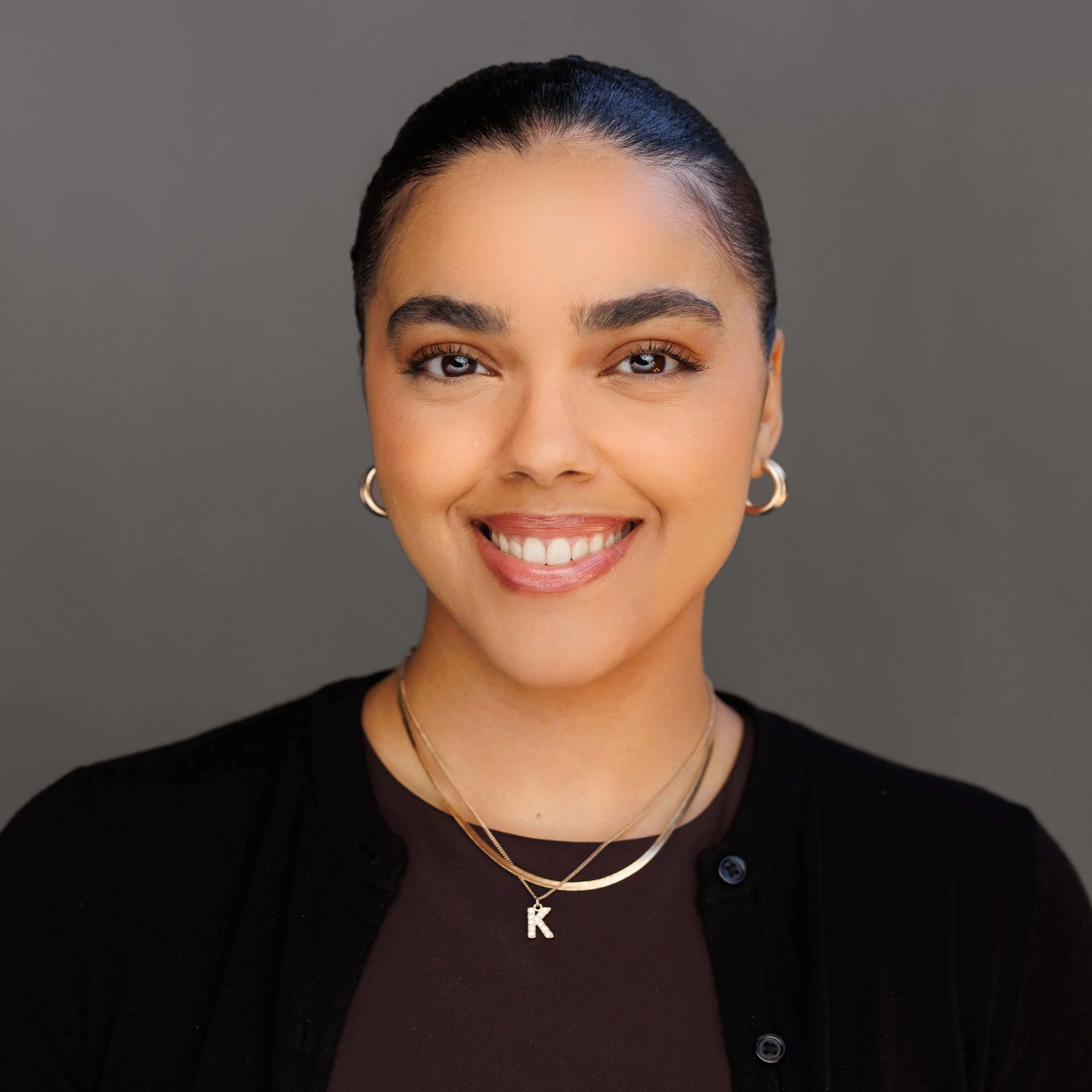 A young woman with dark hair pulled back, smiling warmly, wearing gold hoop earrings, layered gold necklaces including one with the letter 'K', black top, and a black cardigan or jacket, against a neutral gray background.