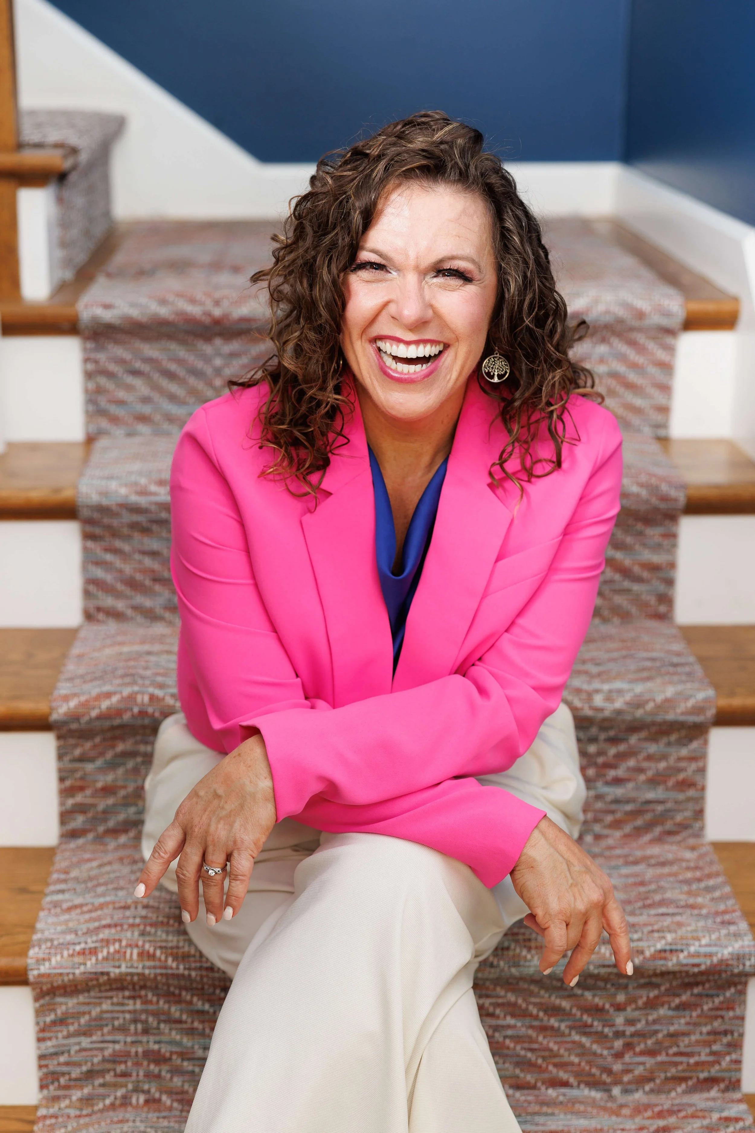 A woman with curly brown hair wearing a bright pink blazer, white pants, and earrings, sitting on a staircase and smiling.