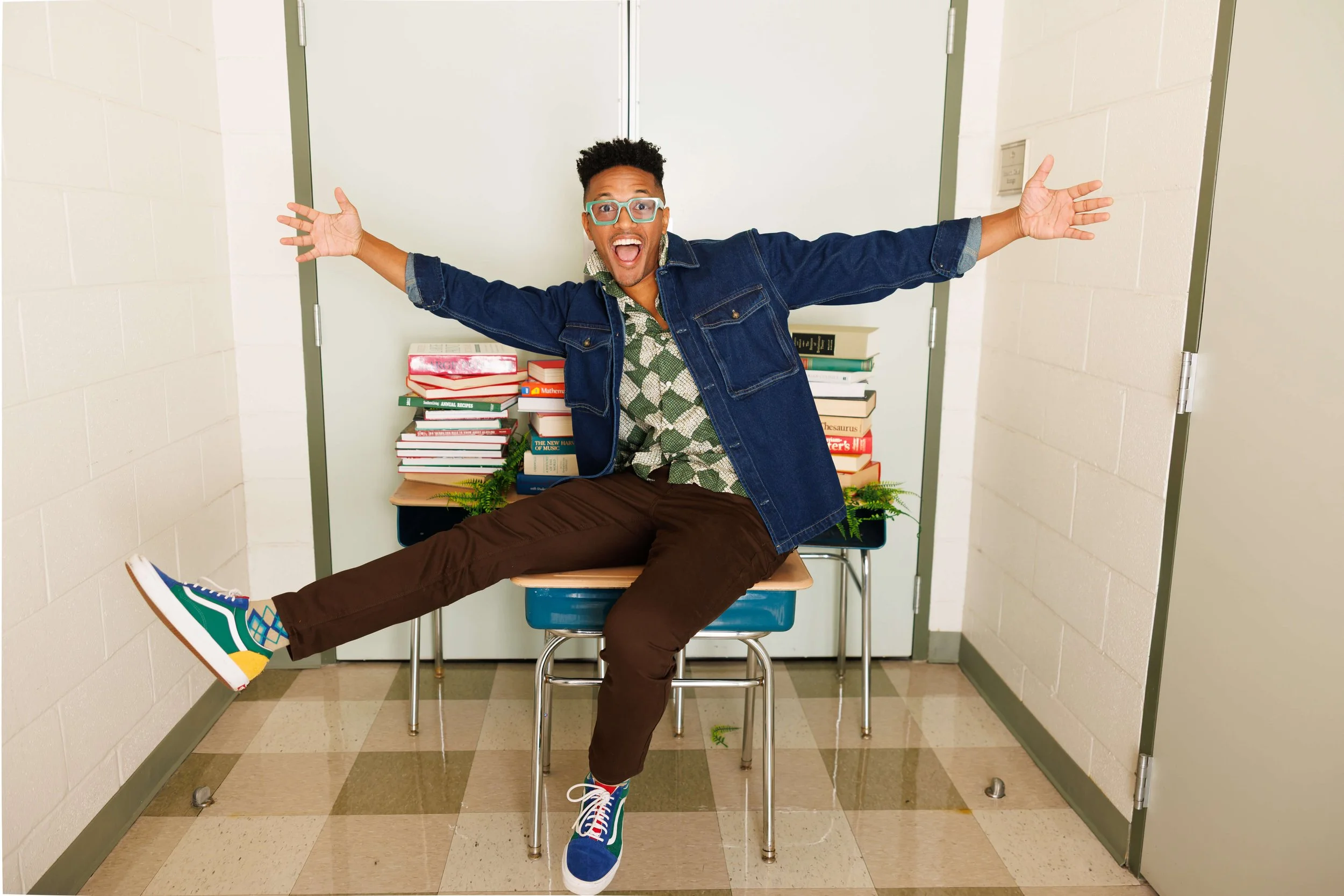 A man with glasses, a denim jacket, a patterned shirt, and colorful sneakers sitting on a desk in a classroom, smiling broadly with arms outstretched.