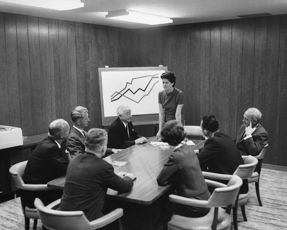 A black and white photo of a woman standing at a conference table addressing six seated men in a boardroom with wood-paneled walls and a chart with upward trending lines on a whiteboard.