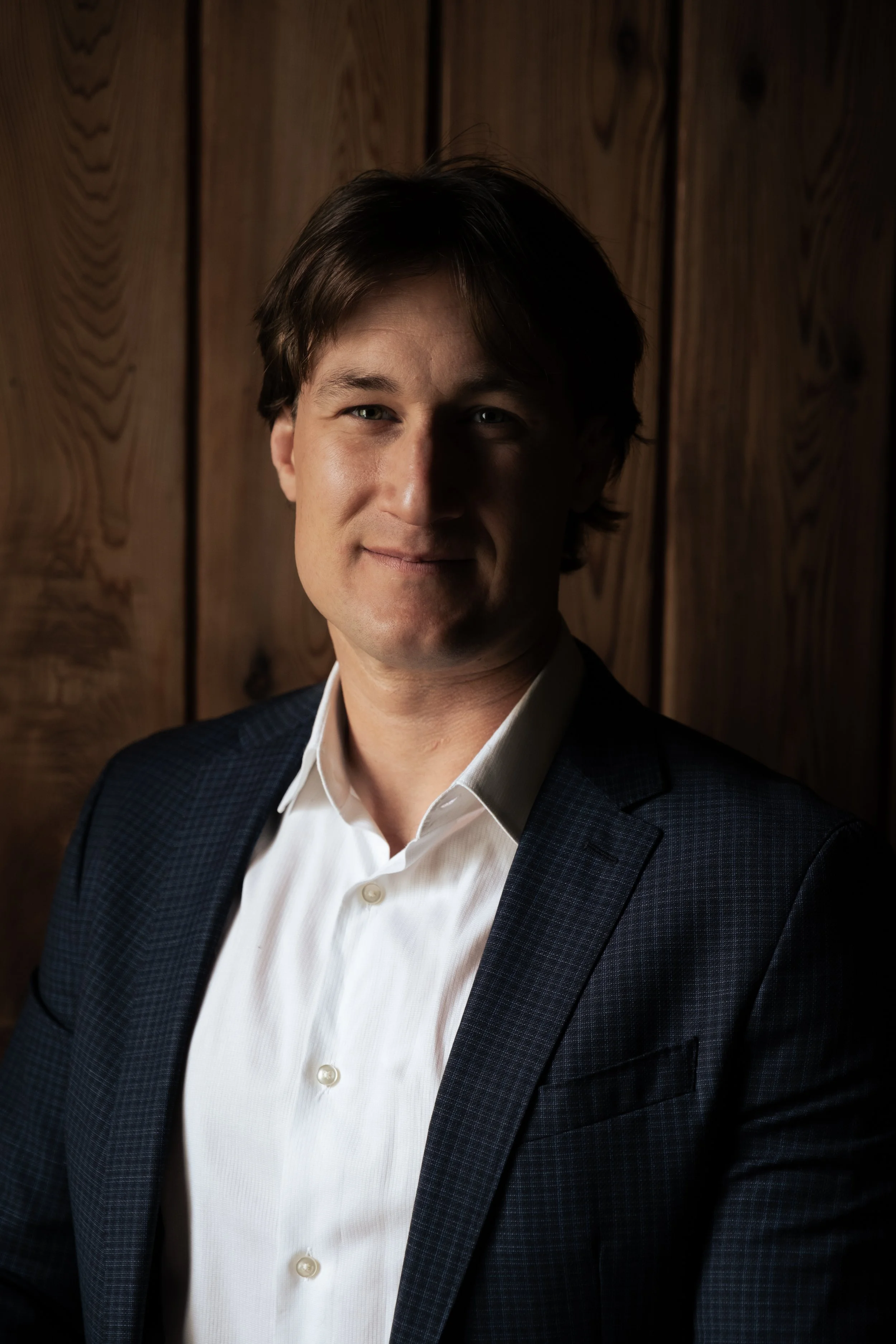 Portrait of a man in a navy checkered suit and white shirt, standing against a wooden panel background with a serious expression.