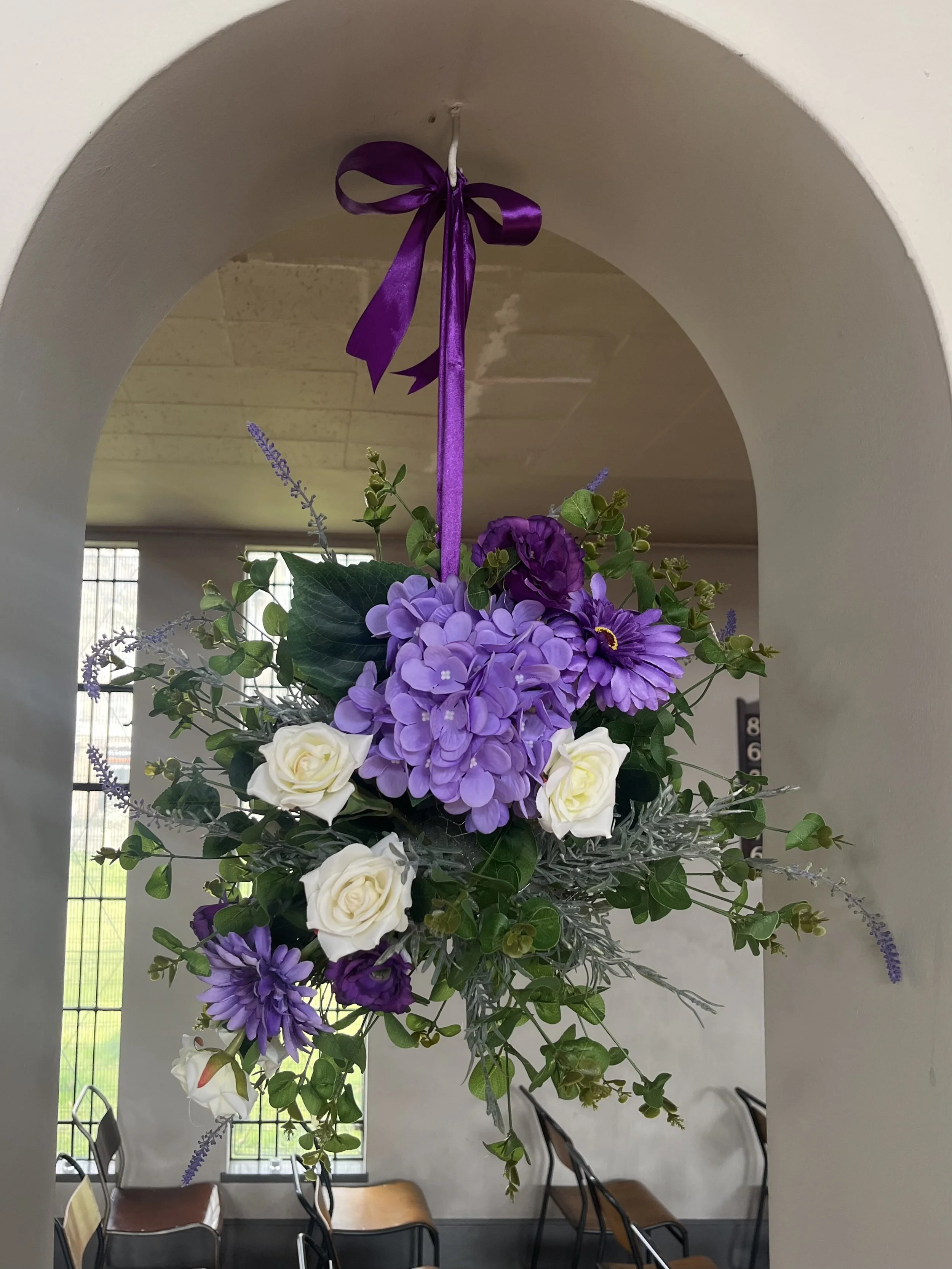 A floral arrangement with purple and white flowers, including hydrangeas, roses, and dahlias, decorated with greenery and hanging from a purple ribbon in an arched indoor setting.