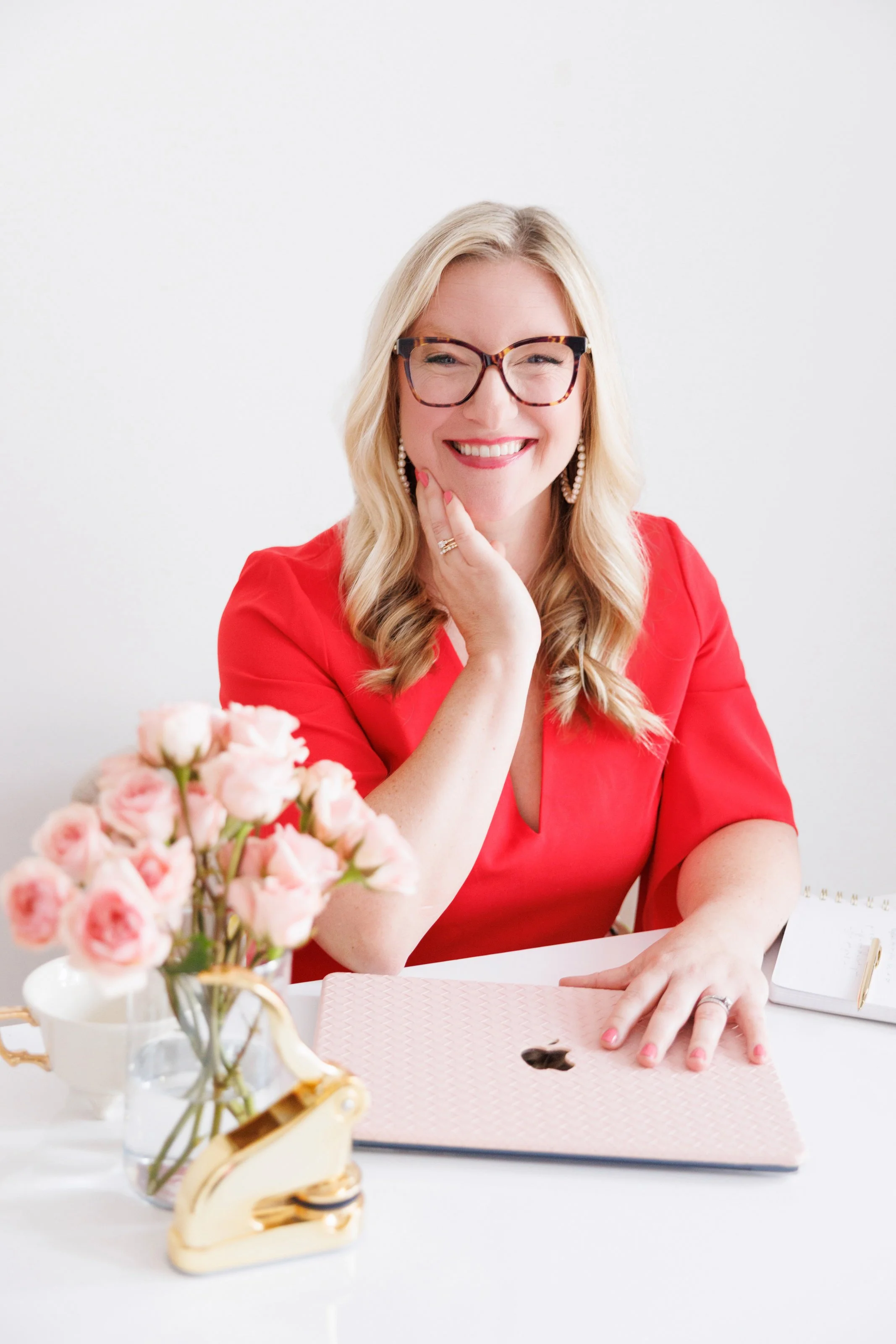 A woman with blonde hair, glasses, and a red dress smiling while sitting at a white table with a laptop, a vase of pink flowers, and a notebook.