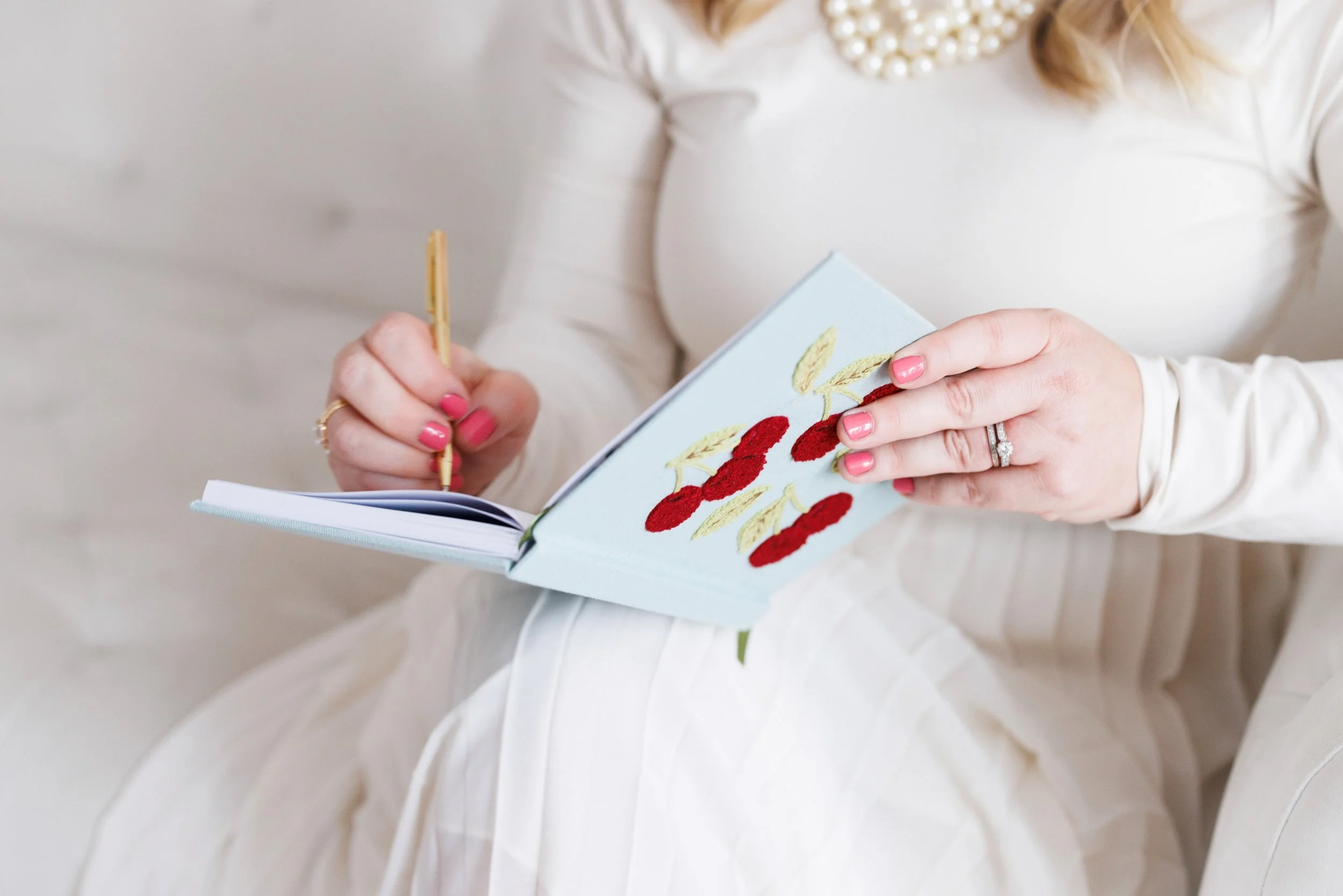 A woman dressed in white, sitting and writing in a small book with embroidered red cherries and green leaves, wearing a pearl necklace and an engagement ring.