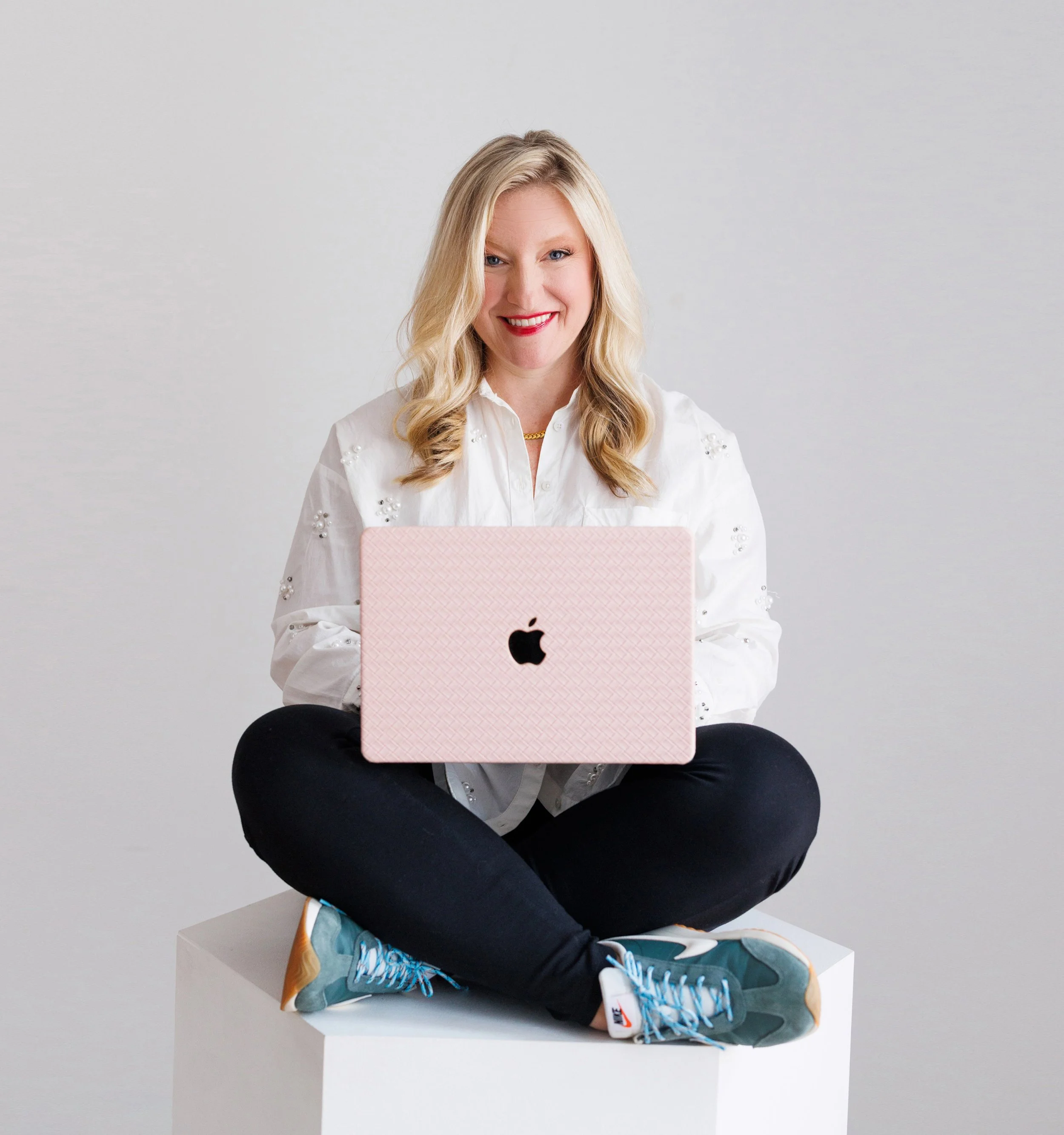 A woman with blonde hair, smiling, sitting cross-legged on a white cube, holding a pink laptop with an Apple logo, dressed in a white blouse, black pants, and sneakers, against a plain white background.