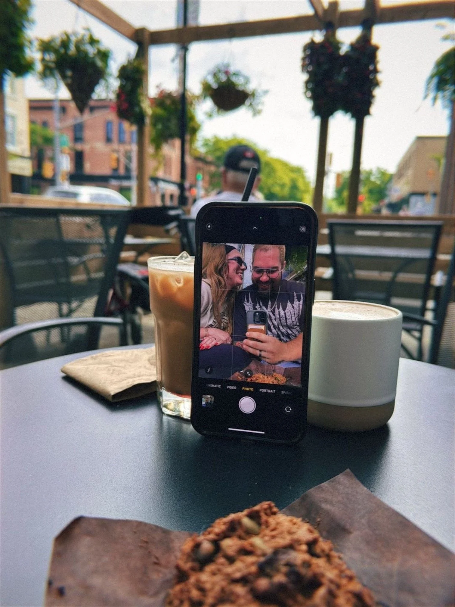 A table at an outdoor cafe with a glass of iced coffee, a bowl of an espresso drink, and a cookie. A smartphone on the table shows a photo of a smiling couple taking a selfie. Hanging flower baskets and buildings are visible in the background.