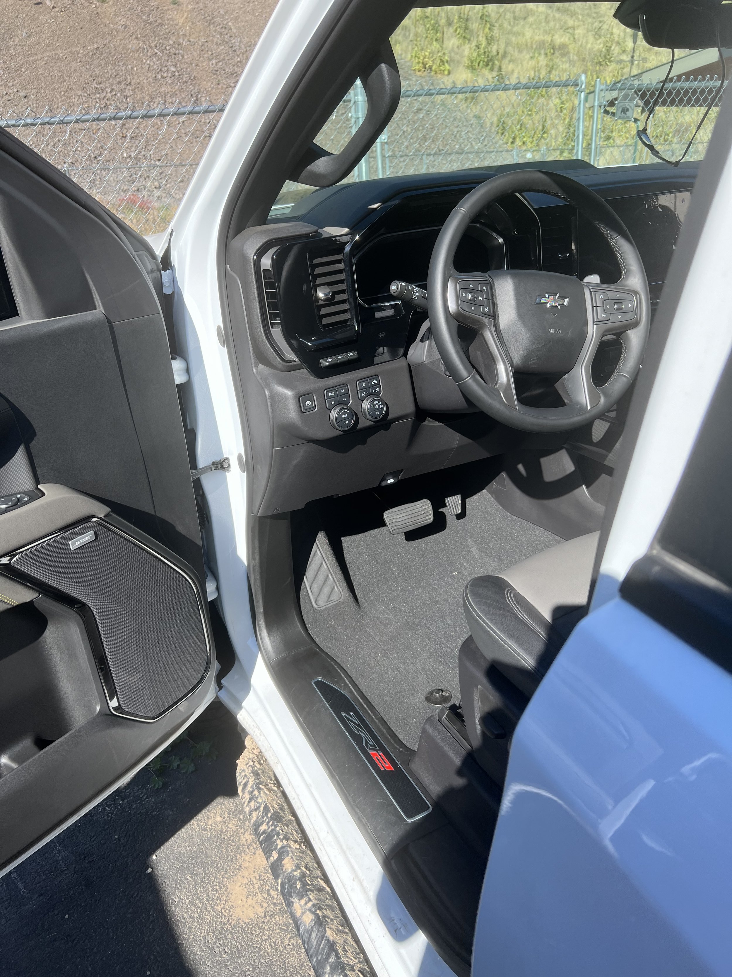 Interior of a white vehicle showing the driver's side door open, steering wheel, dashboard controls, pedals, and "ZR2" marked step.