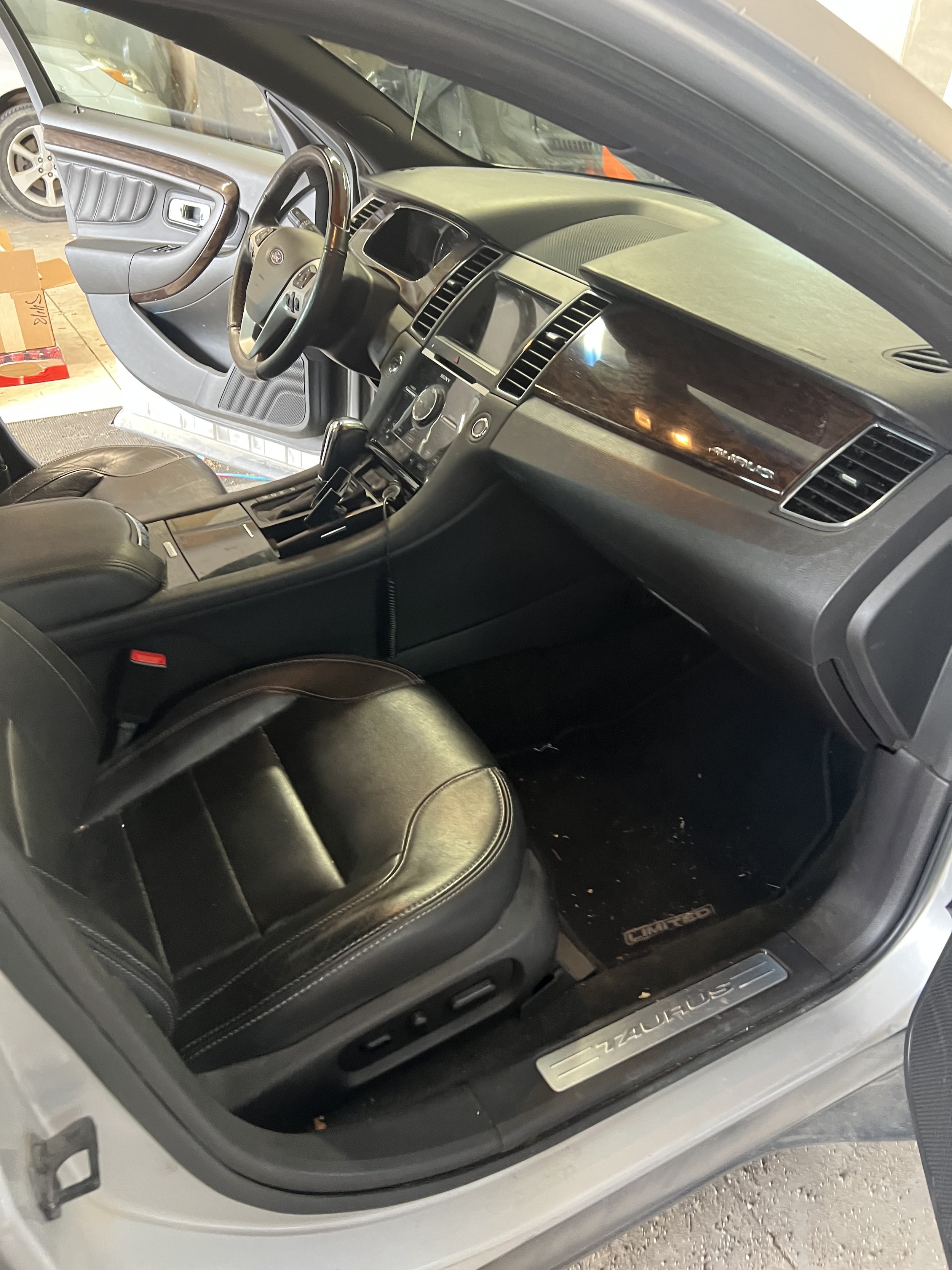 Interior of a car with black leather seats, wood trim on the dashboard and door panel, and a visible steering wheel. The car door is open, and the interior appears clean and well-maintained.