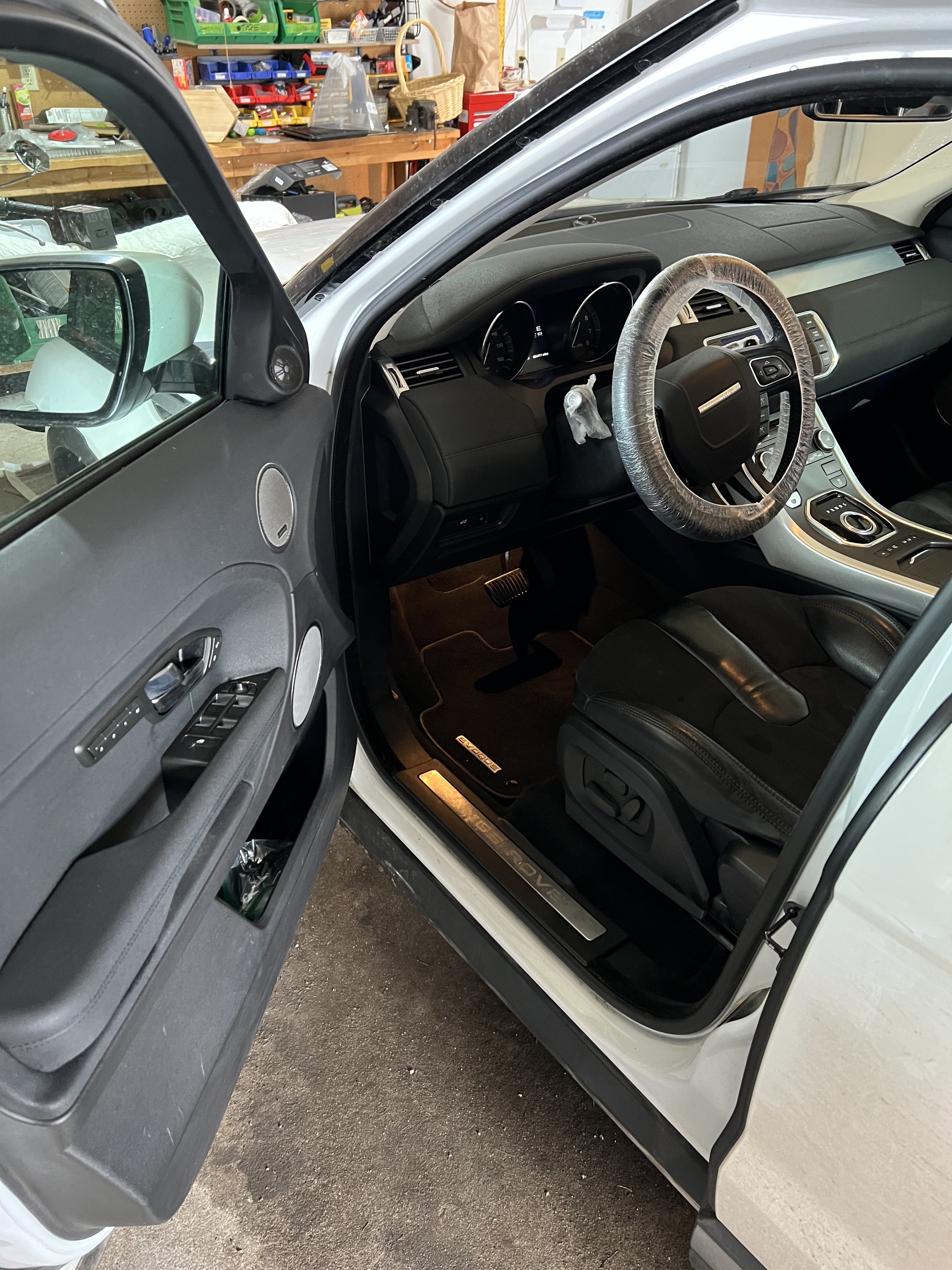 Interior of a modern car with open driver's door, showing the dashboard, steering wheel covered with plastic, and black leather seat. The door features control buttons, a speaker, and a storage compartment. The floor mat has a visible brand label.