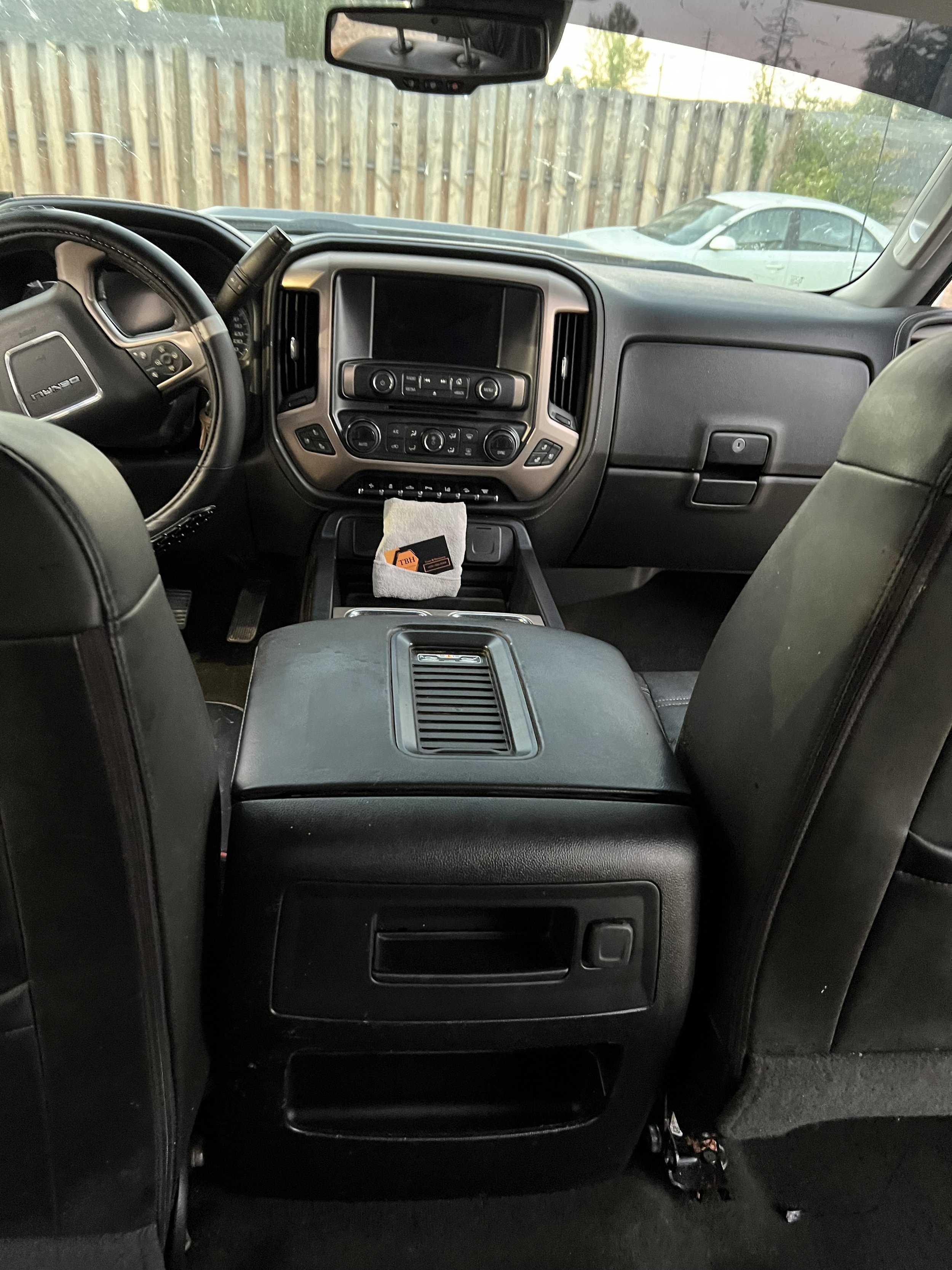 Interior of a vehicle showing the dashboard, steering wheel, and center console with a fast-food bag on it. A wooden fence and a parked car are visible through the rear window.
