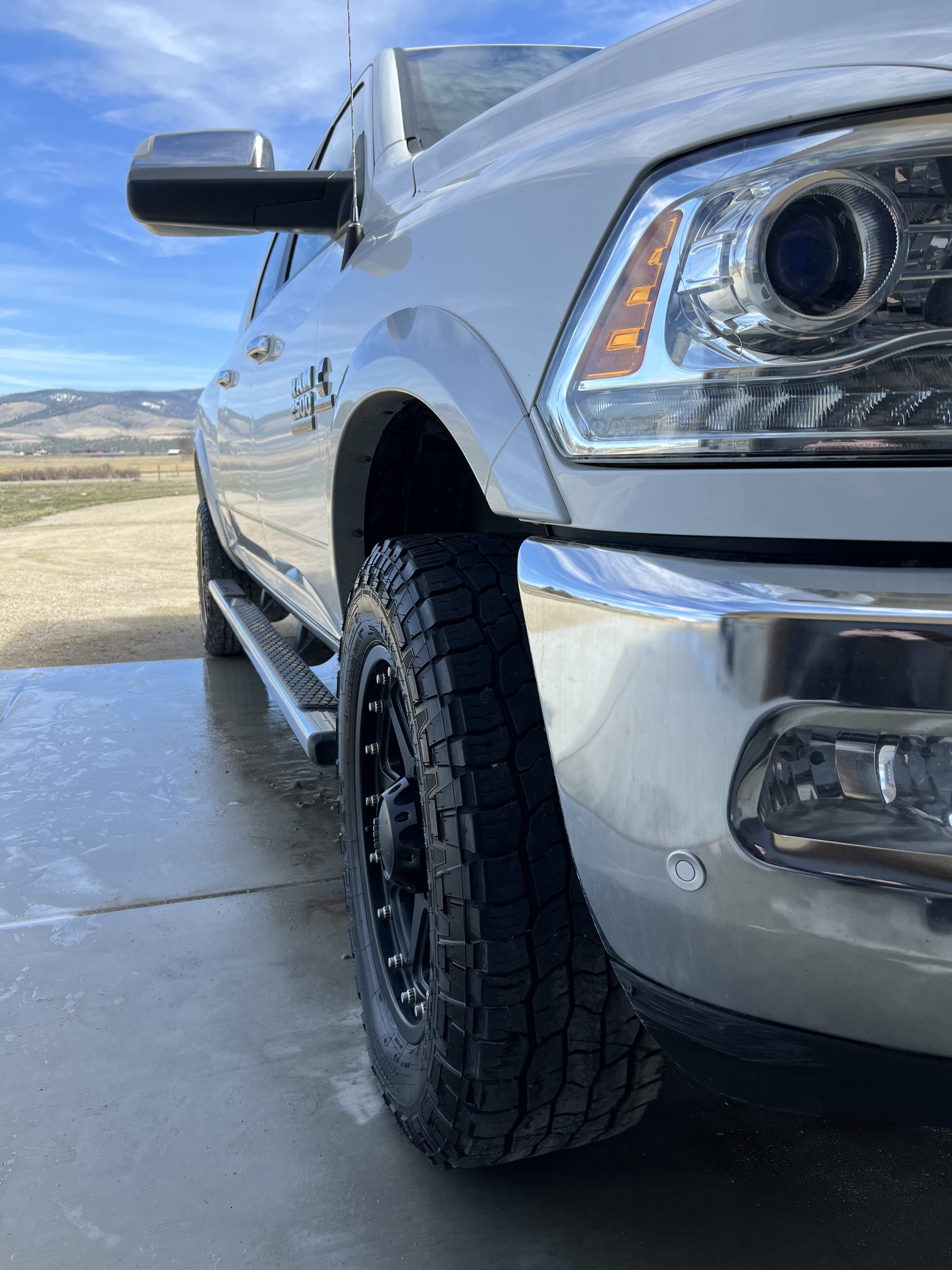 Front end of a white pickup truck with black wheels parked on a concrete driveway, mountains in the background.