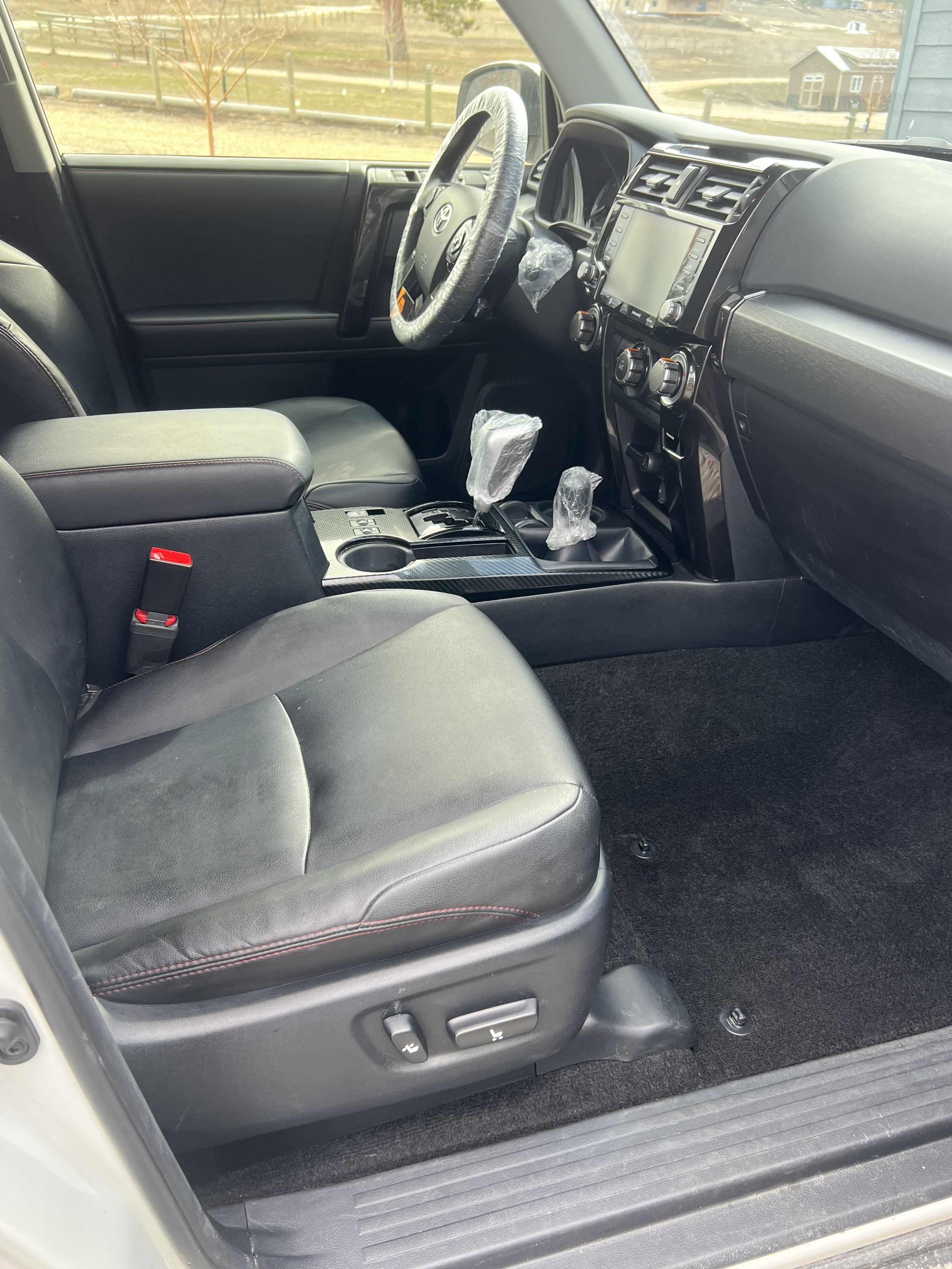 Interior of a car showing leather seats, dashboard with control knobs, gear shift, and steering wheel covered in protective plastic, parked in an outdoor setting.
