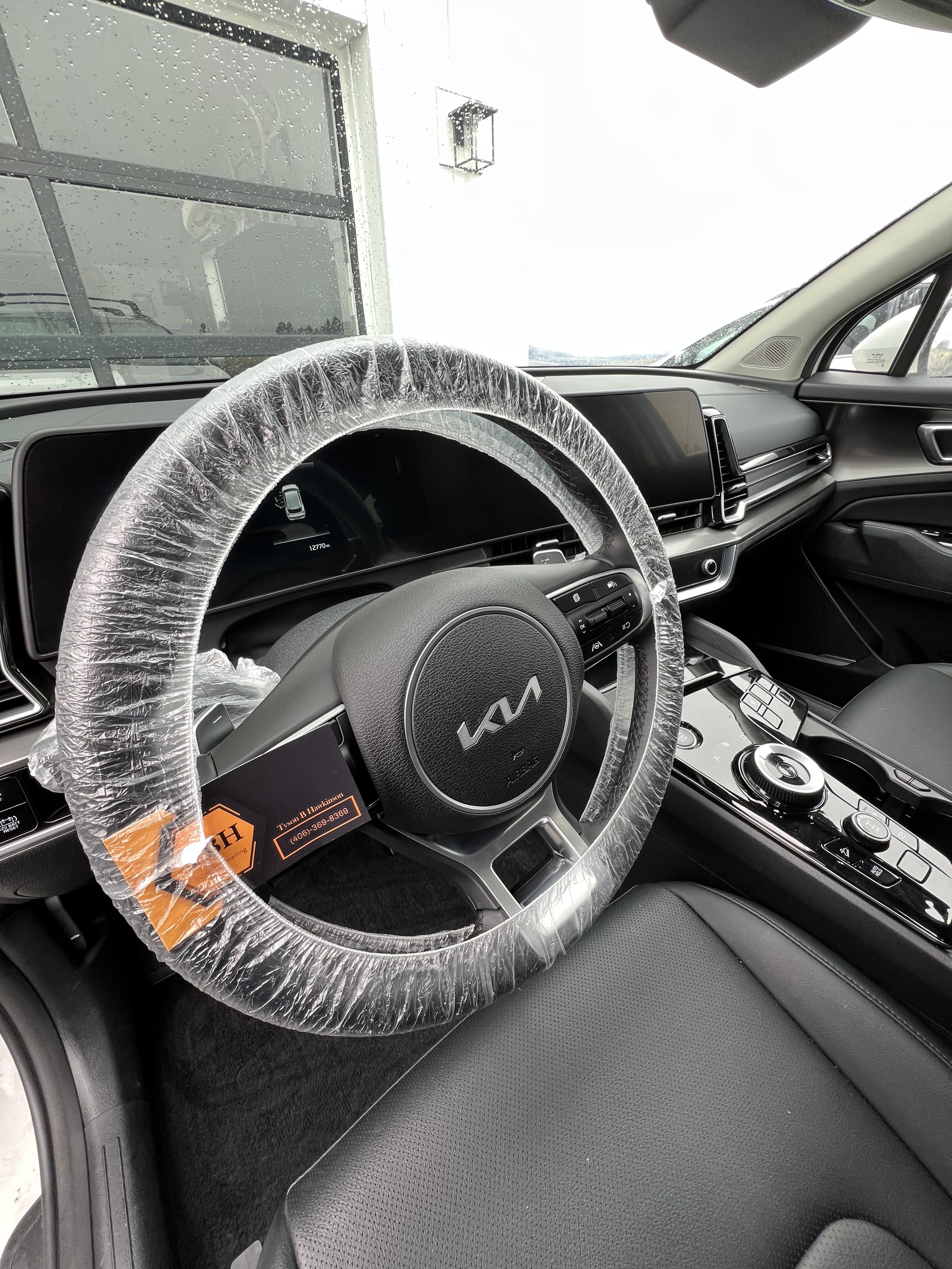 Interior of a new car showing a wrapped steering wheel with a Kia logo, alongside a central console with control buttons and a gear selector.