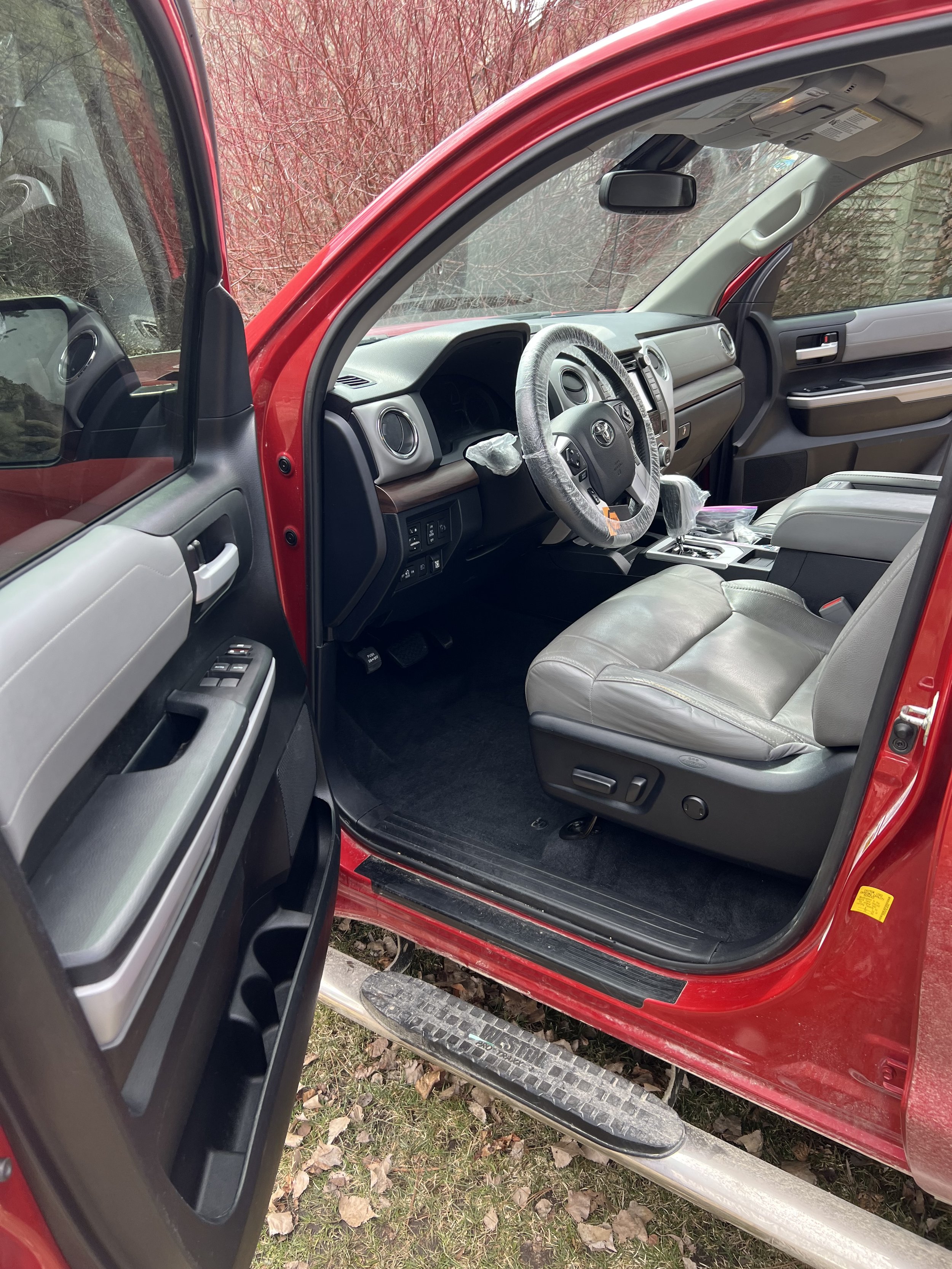 Interior view of a red pickup truck with open driver-side door, showing gray leather seats and a steering wheel covered in plastic, indicating a new or recently cleaned vehicle.