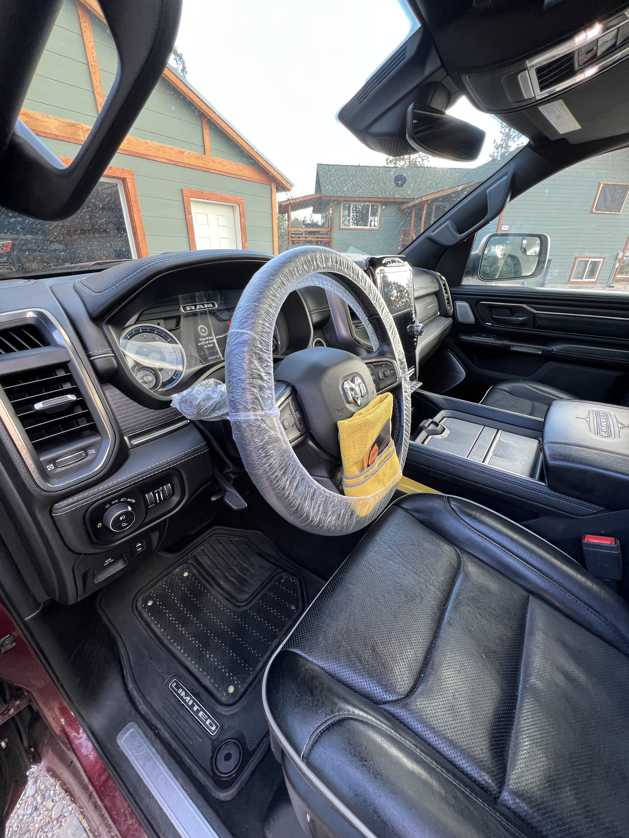 Interior of a RAM truck showing a steering wheel with plastic protection, dashboard, black leather seats, and a view of the garage outside.