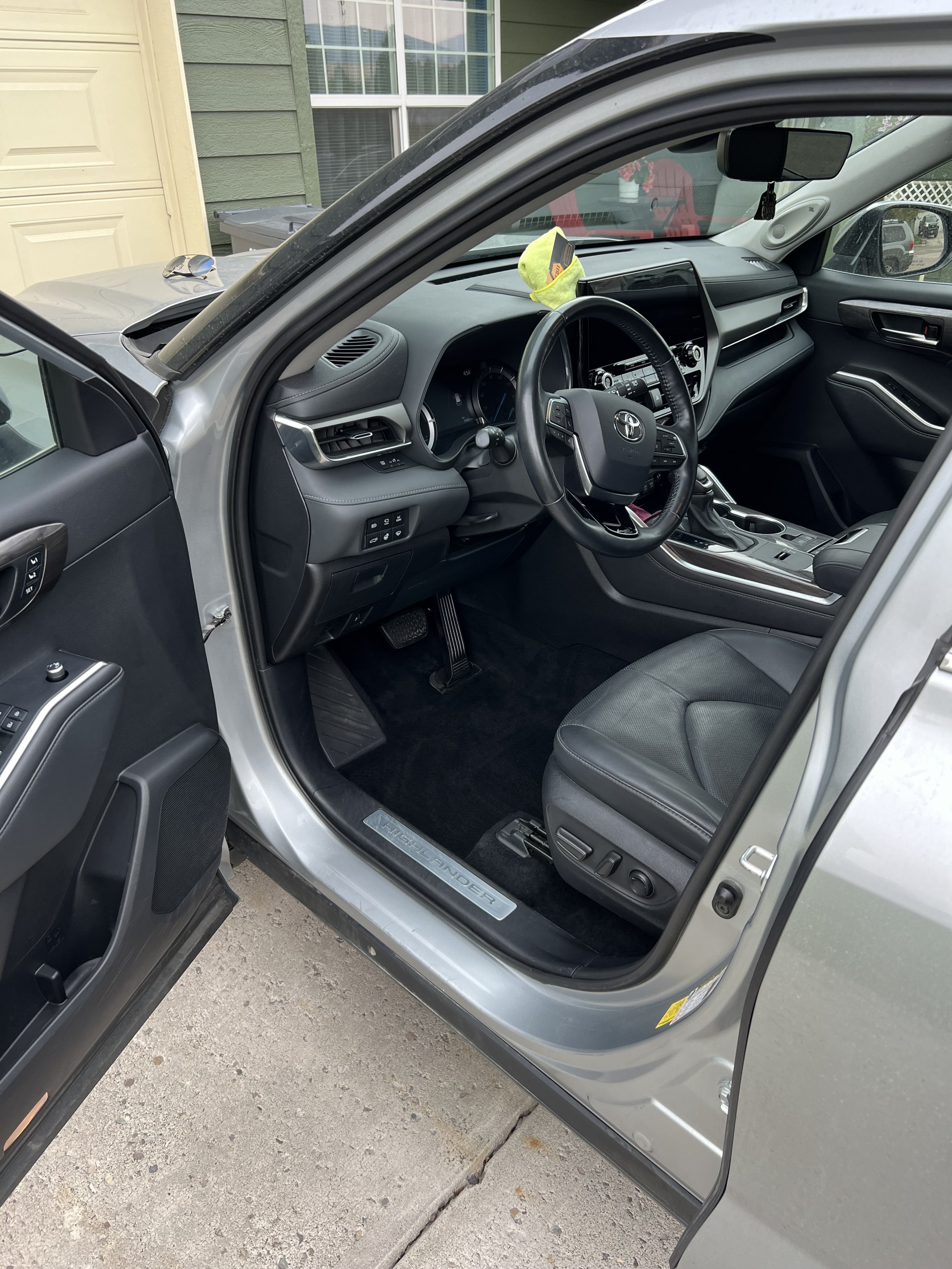 Interior of a parked car with an open driver's door showing a steering wheel and dashboard, leather seats, and a hanging air freshener.