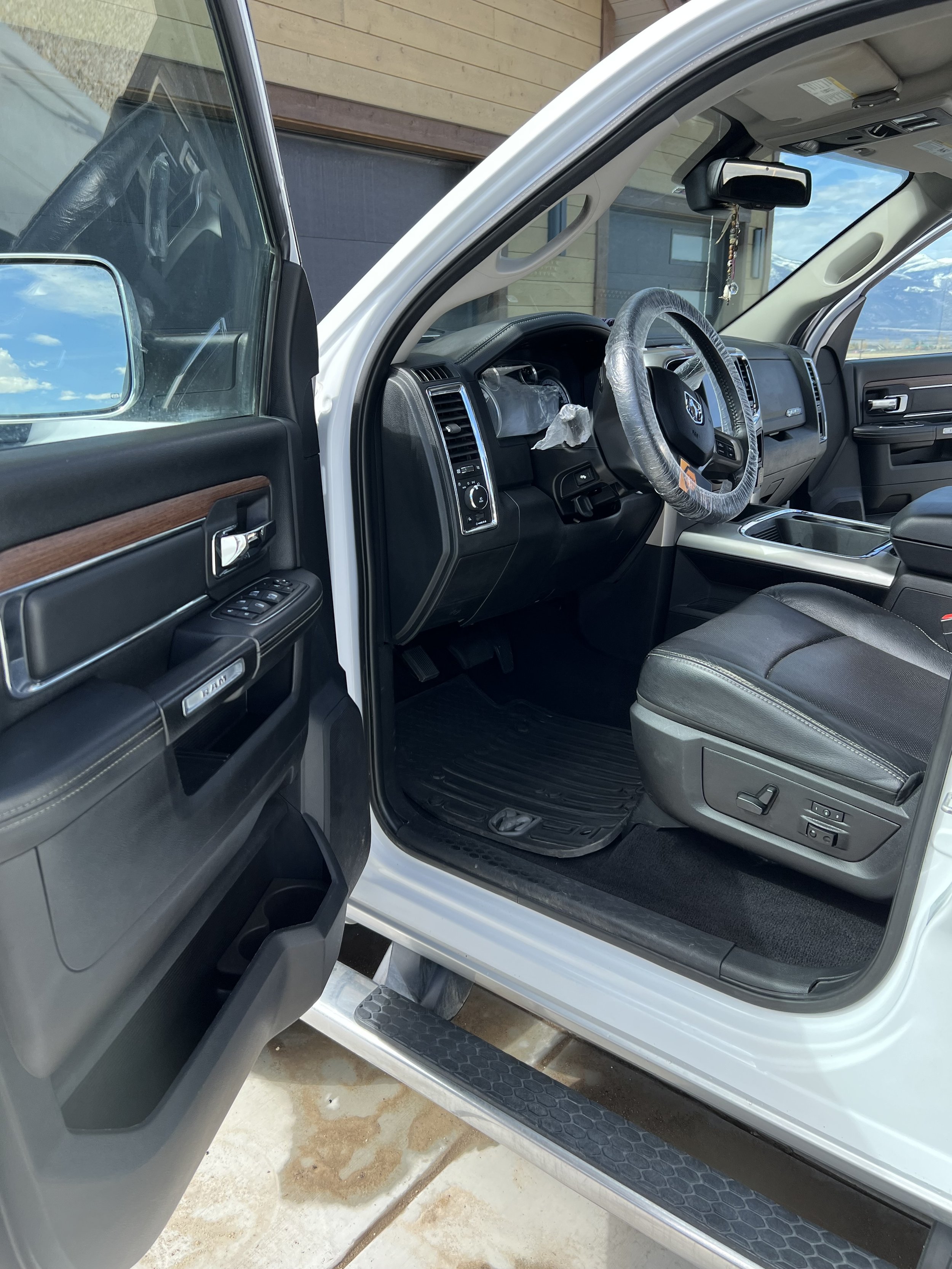 Interior view of a white truck's driver's side, showing the steering wheel with a cover, dashboard, seat controls, and the open door with a visible side mirror.