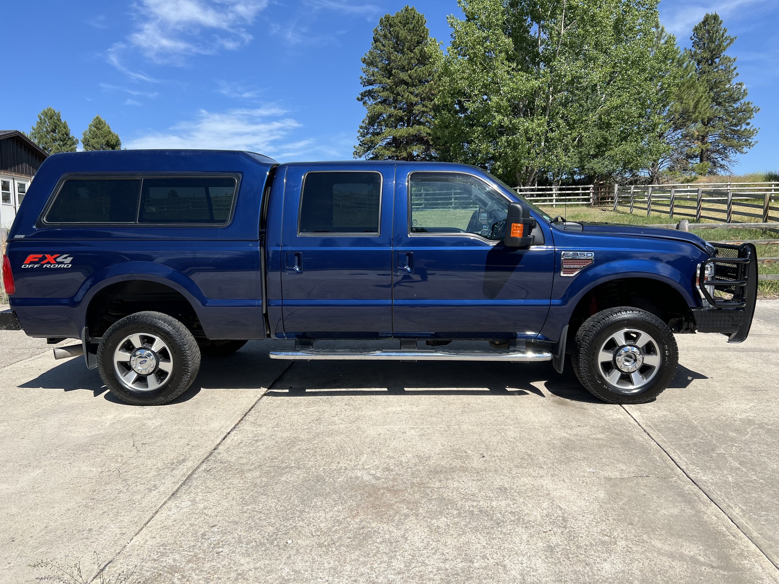 Blue pickup truck with a camper shell, parked on a concrete driveway, surrounded by trees and a fence.