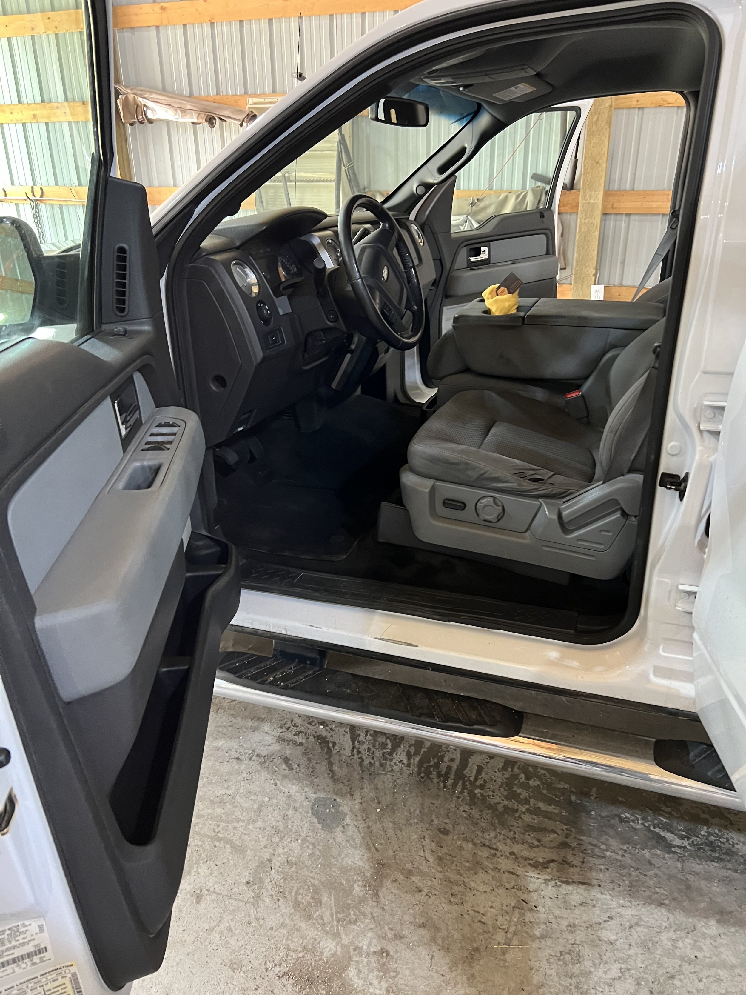 Interior of a white pickup truck with open driver's side door, showing the steering wheel, dashboard, gray seat, and door panel.
