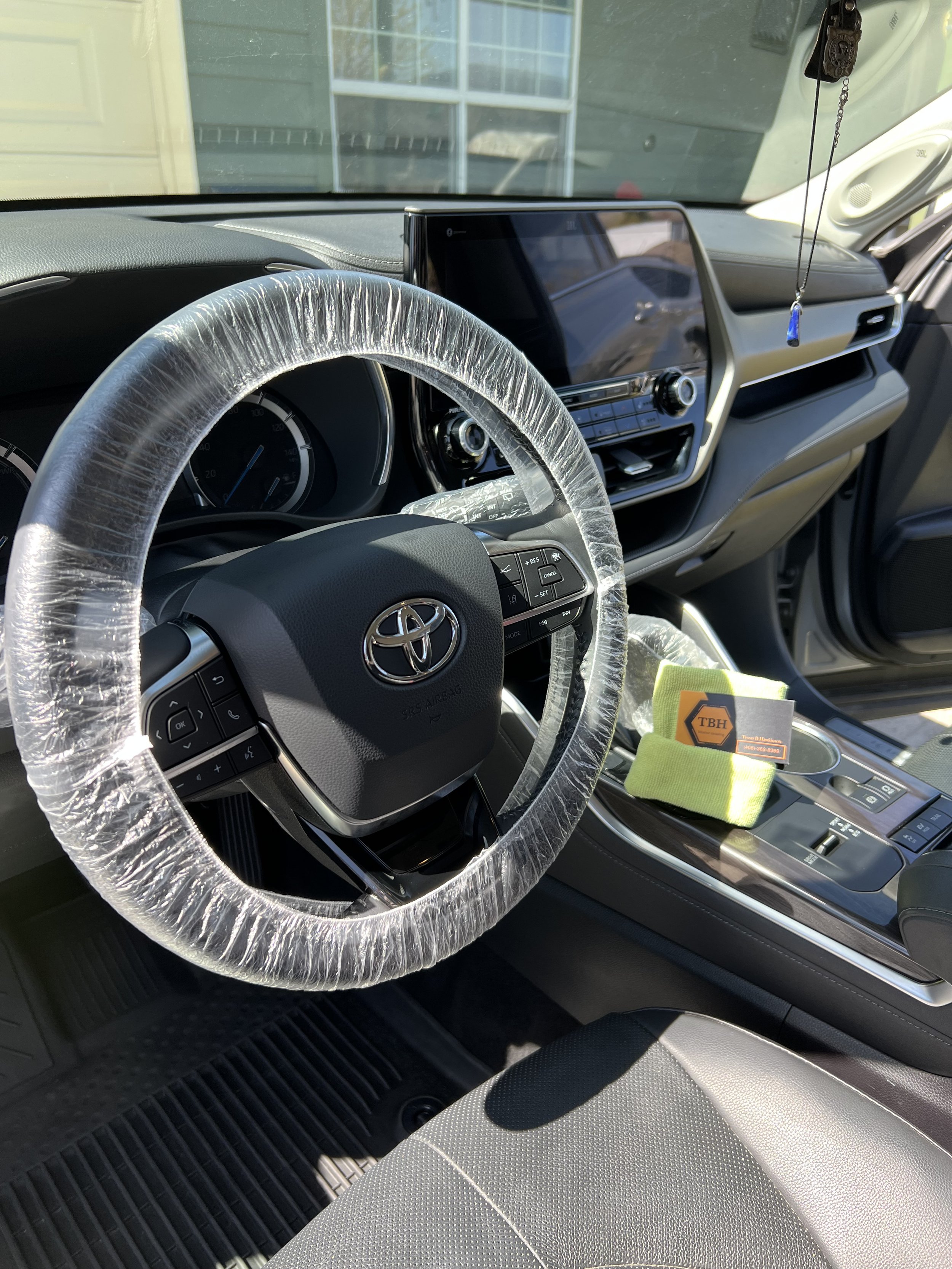 Interior of a Toyota vehicle showing a plastic-wrapped steering wheel and dashboard, infotainment display, rubber floor mats, and a microfiber cloth on the center console.