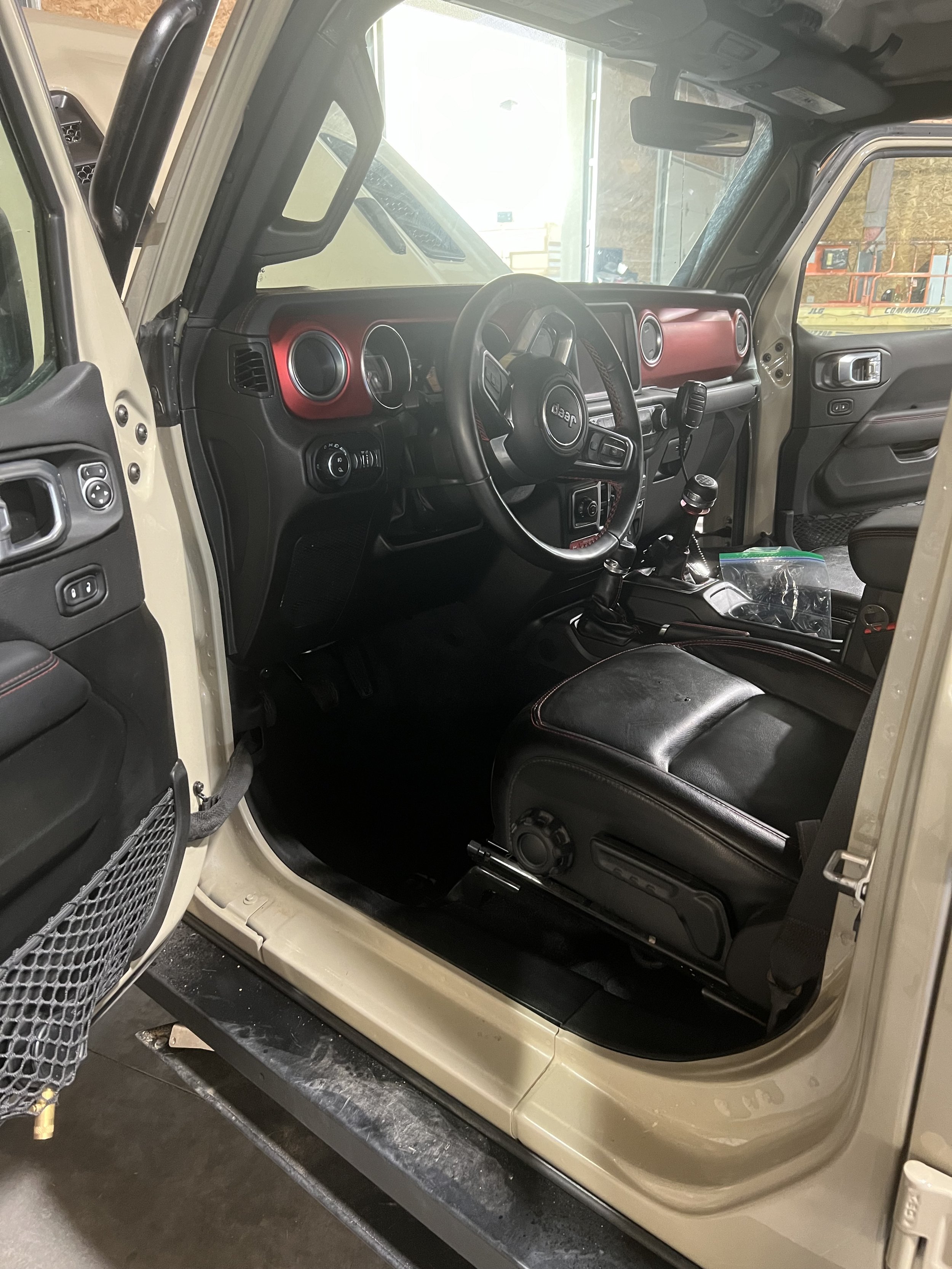 Interior of a Jeep with open driver door, featuring leather seats, red dashboard accents, steering wheel, and manual gear shift.