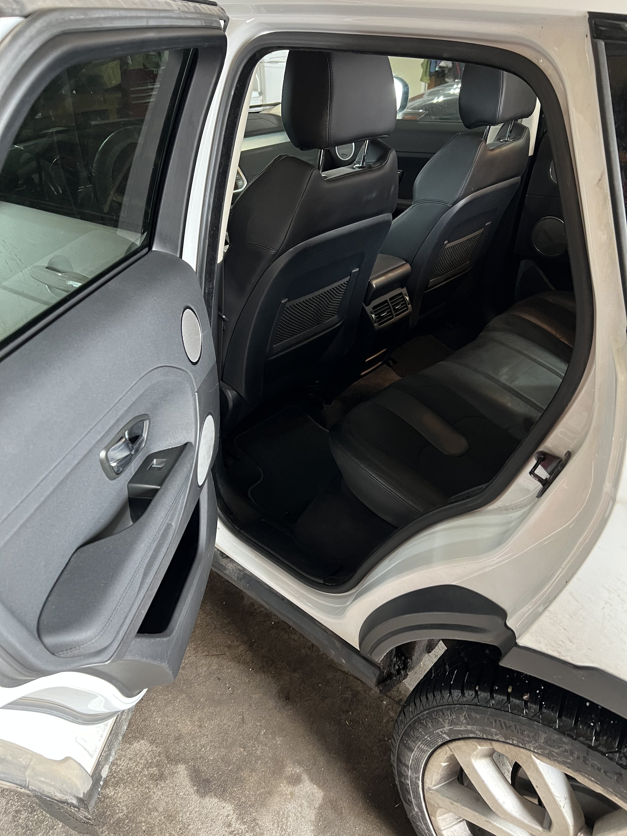 Interior view of a car's back seat with an open rear door, showing black leather seats and armrest, with visible door panel and tire.