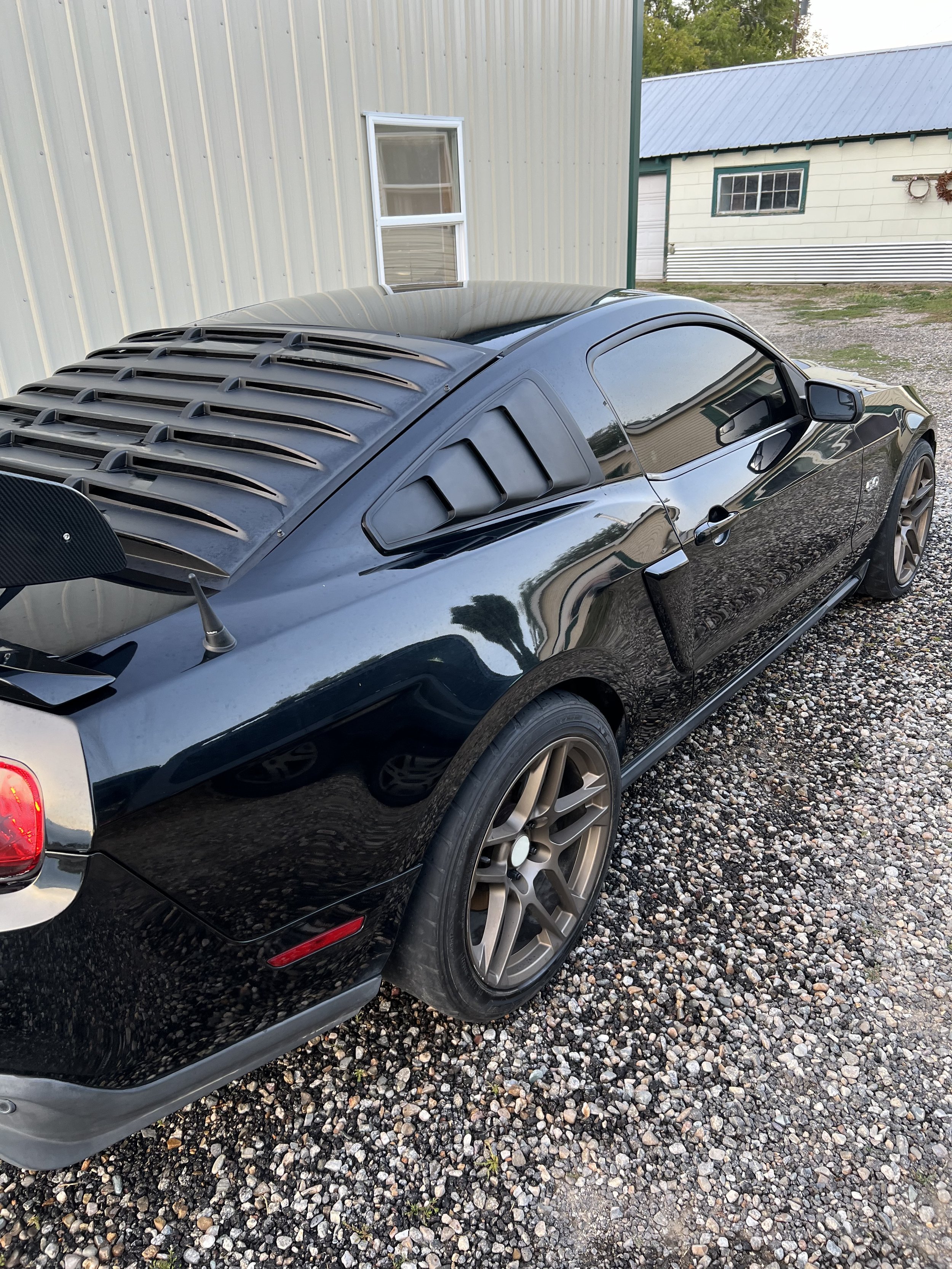 Black sports car with louvered rear window and aftermarket spoiler parked on gravel near a metal building.
