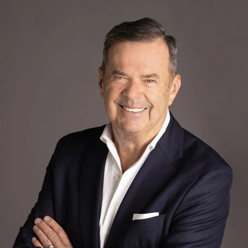 Martin Wiedmann in a white shirt and dark blazer smiling with arms crossed against a gray background.