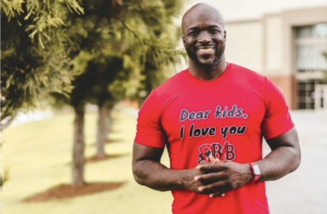 Ricky Sapp standing outdoors in front of trees, wearing a red t-shirt that says "Dear kids, I love you" and has a logo related to a fraternity or organization.