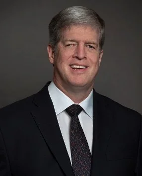 Dr. Kirk Adams in a dark suit, white shirt, and dark tie smiling against a dark background.