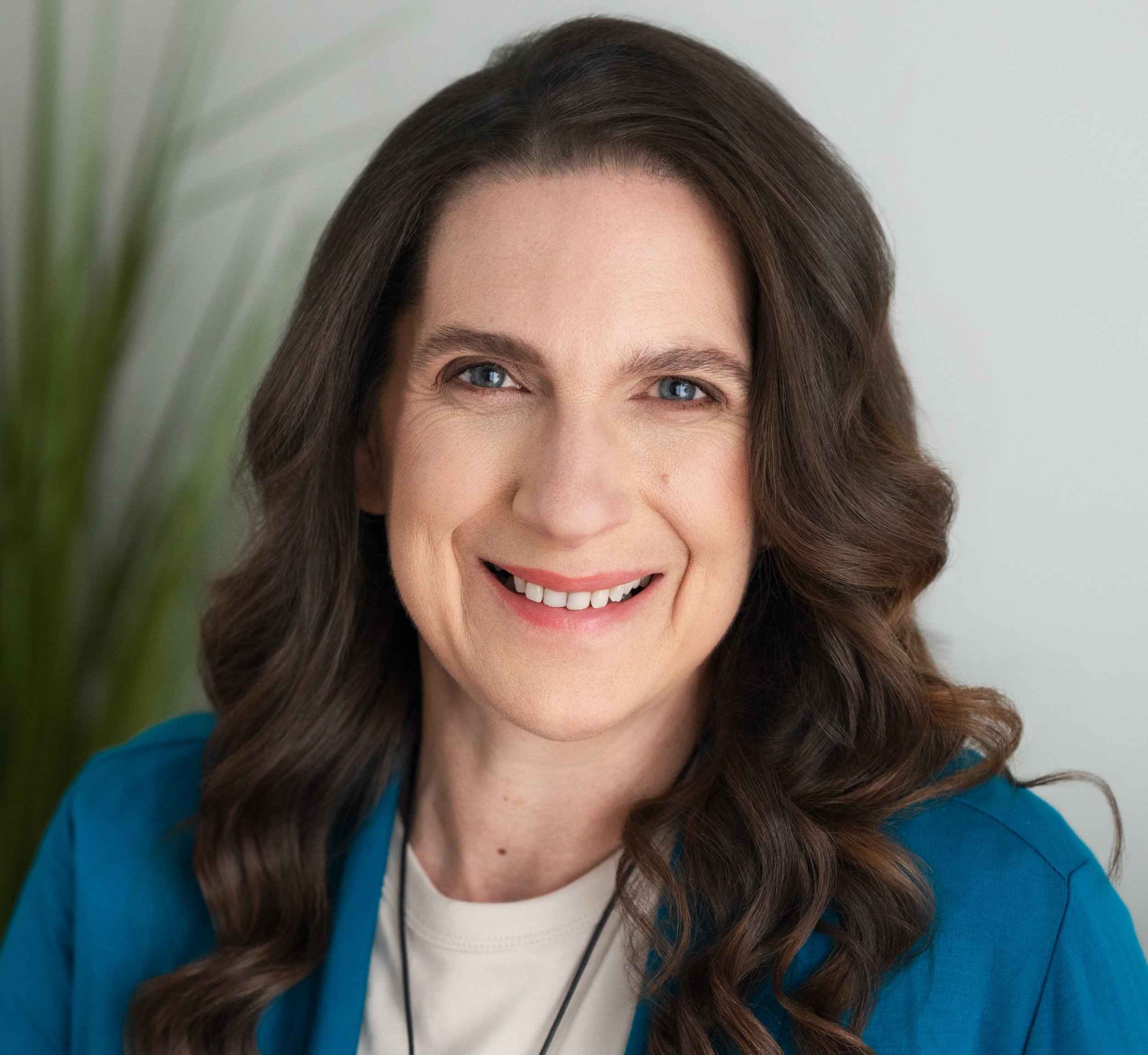 A woman with long brown hair, wearing a teal top, smiling at the camera against a plain white background.