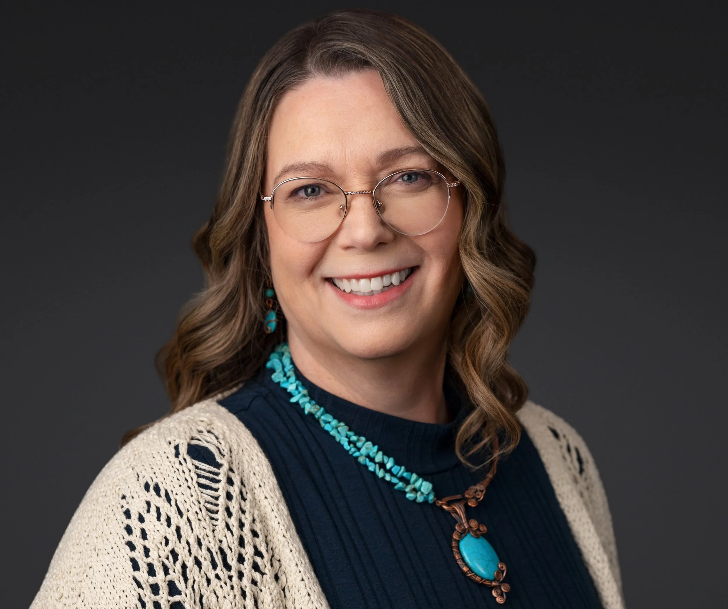 Smiling woman with shoulder-length blonde hair and glasses, wearing a denim shirt and beaded necklace, outdoors with a body of water and a distant shoreline in the background.