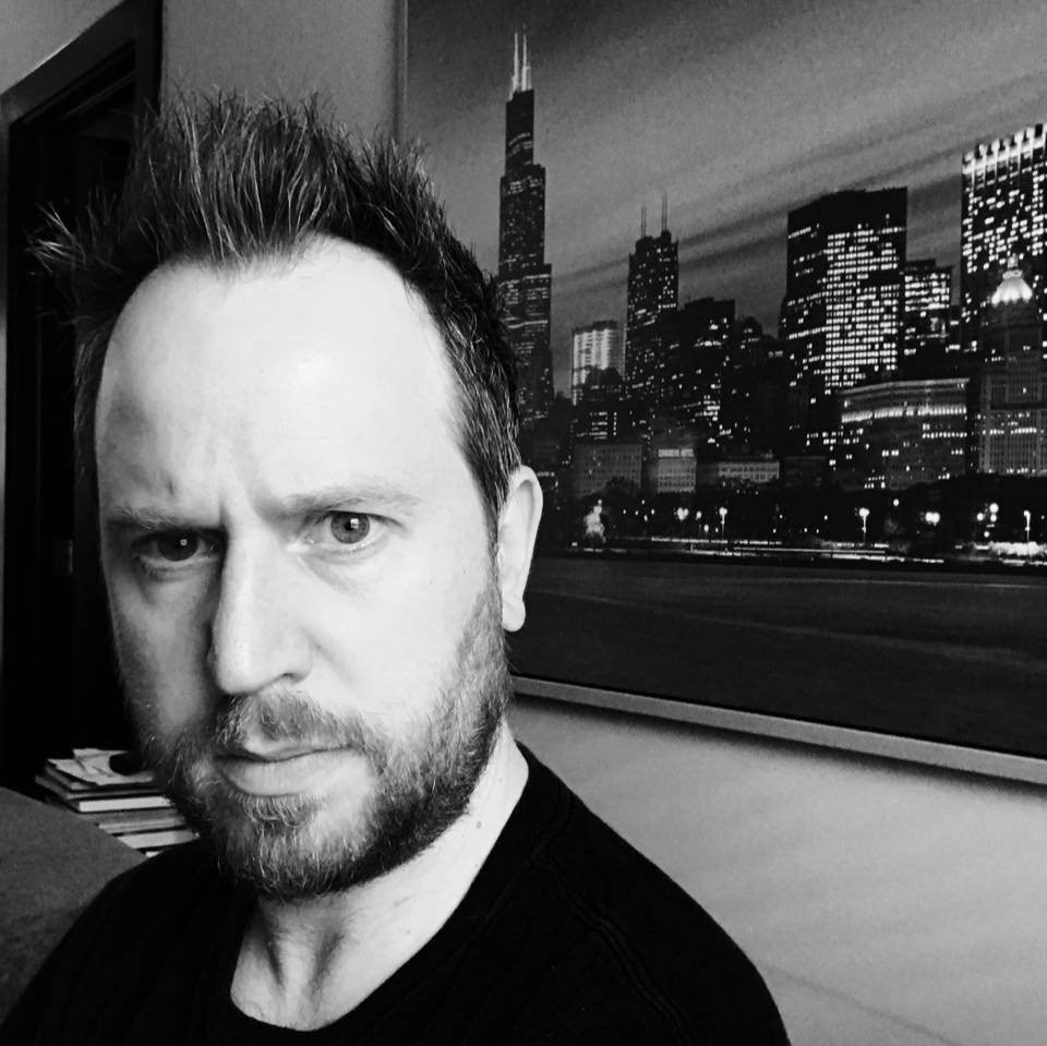 Black and white photo of author Grant Piercy, a bearded man with spiked hair in a black shirt in front of a portrait of the Chicago skyline.
