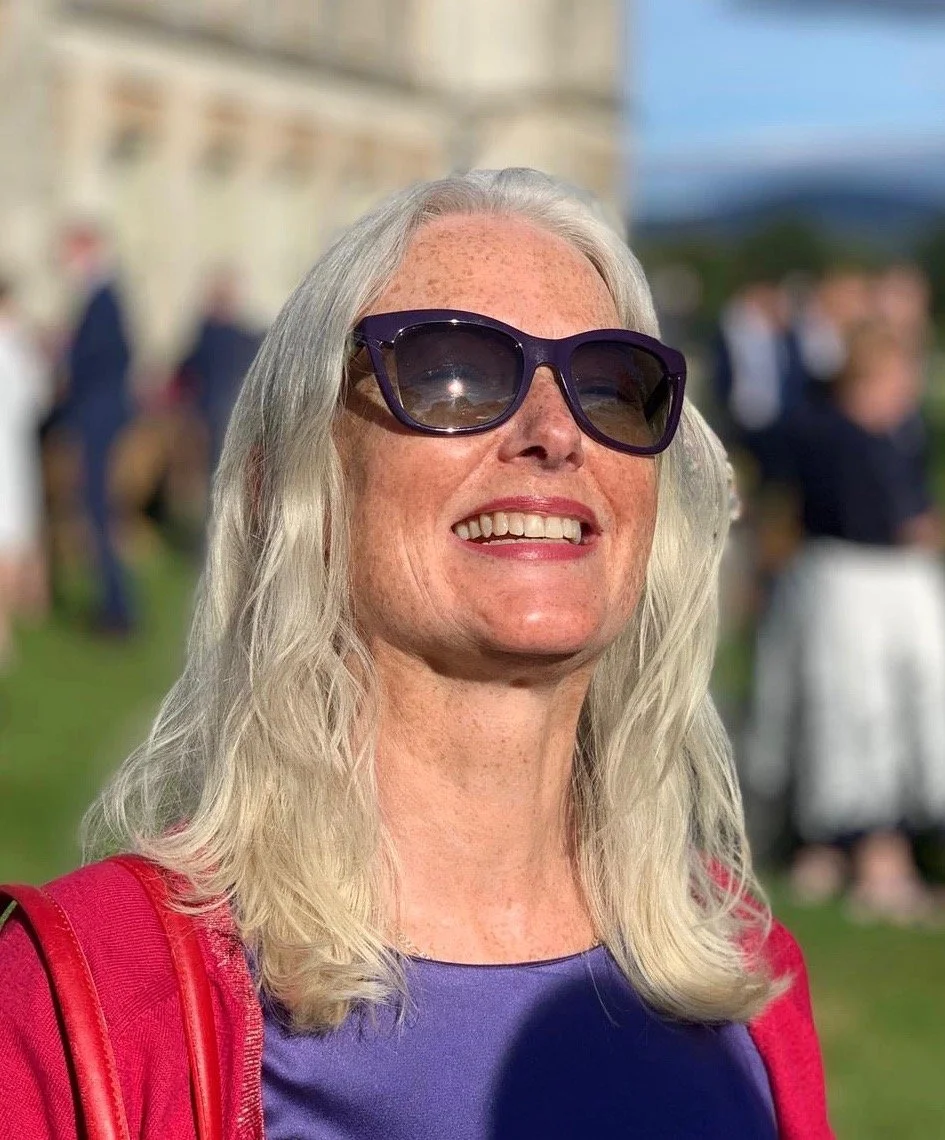 A smiling woman with gray hair wearing sunglasses and a red jacket outdoors, with a blurred background of people and a building.
