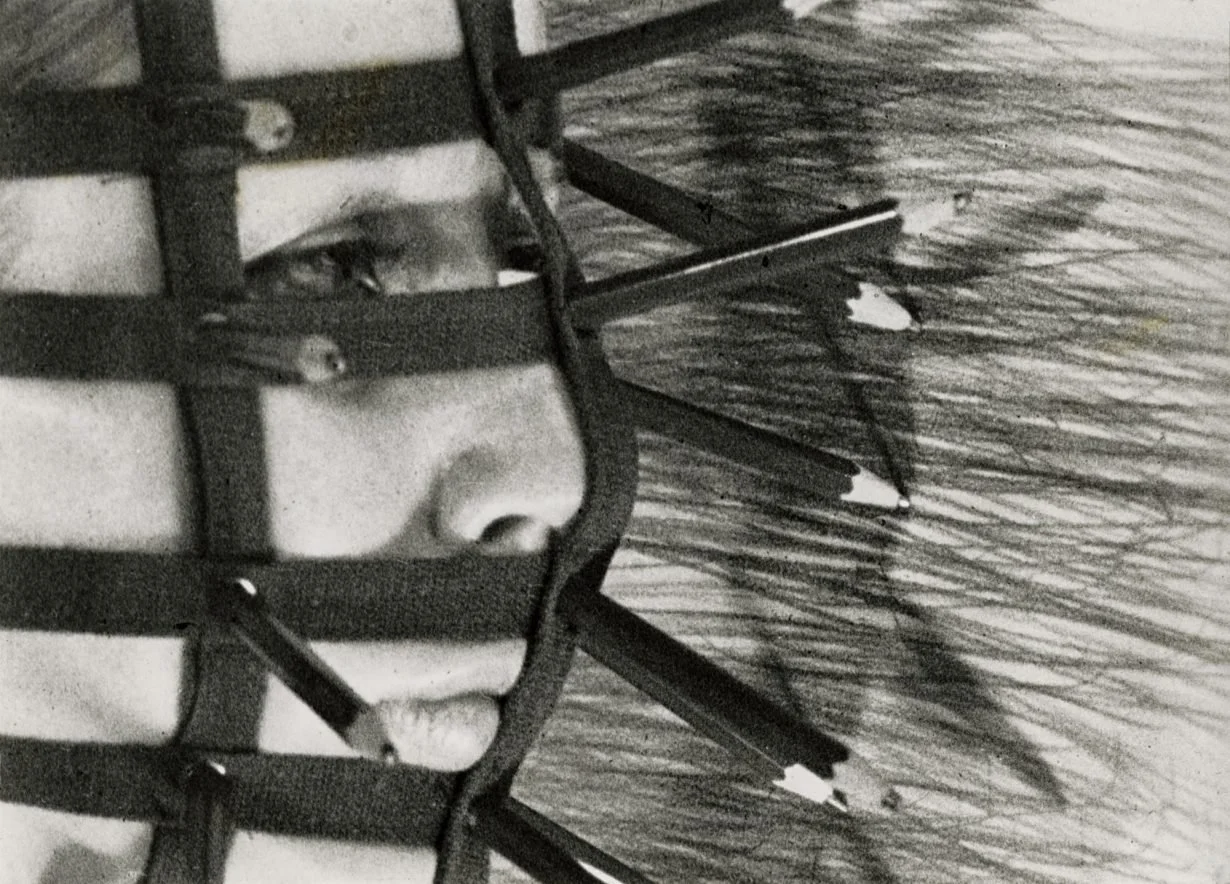 A close-up black and white photo of a person's face with goggles and a harness, possibly an aviator or pilot, with water and sky in the background.