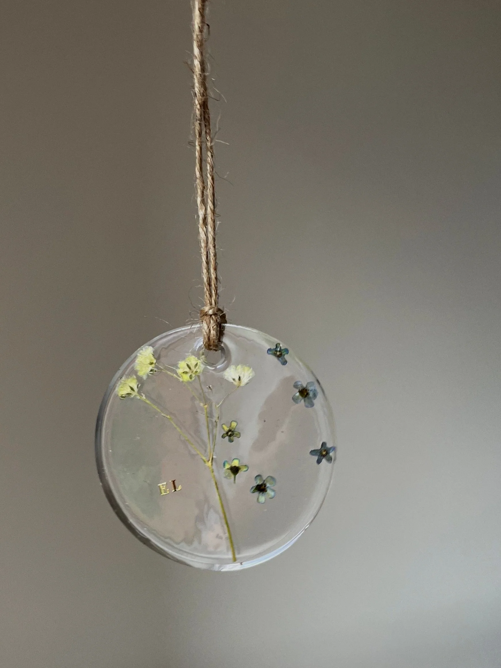 A round, transparent ornament with small yellow flowers and blue flowers embedded inside, hanging from a rustic twine string against a plain background.