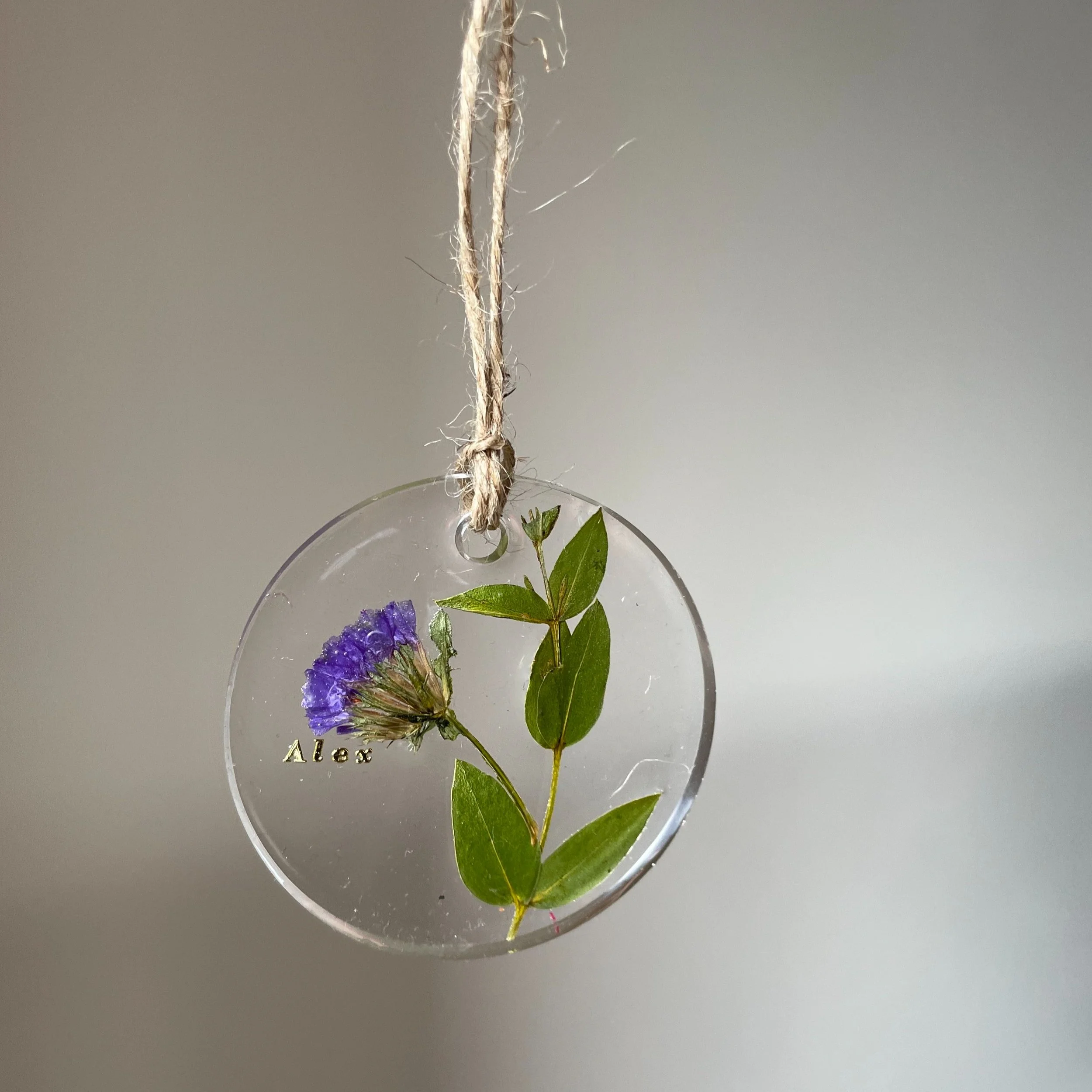 A round glass ornament with purple flower and green leaves inside, hanging from a piece of twine. The name Alex is engraved on the glass.