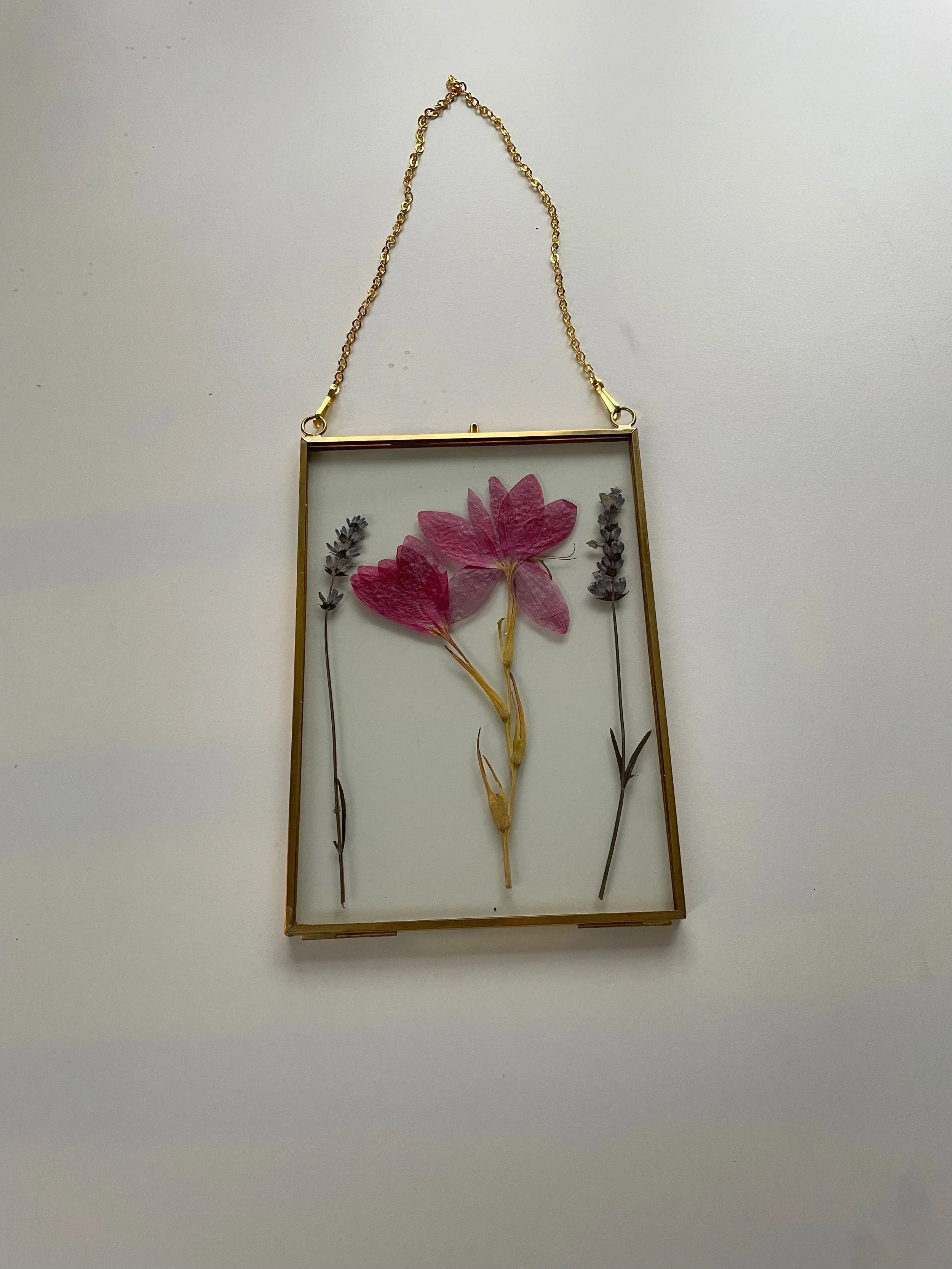 A gold-framed necklace containing pressed pink and purple flowers, hanging from a gold chain.
