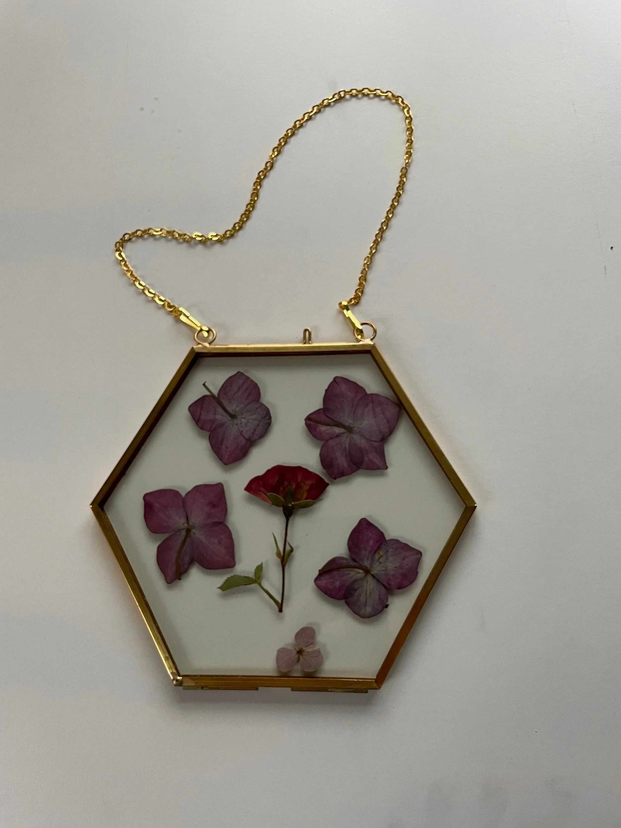 A hexagonal glass pendant necklace with pressed purple and pink flowers inside, attached to a gold chain.