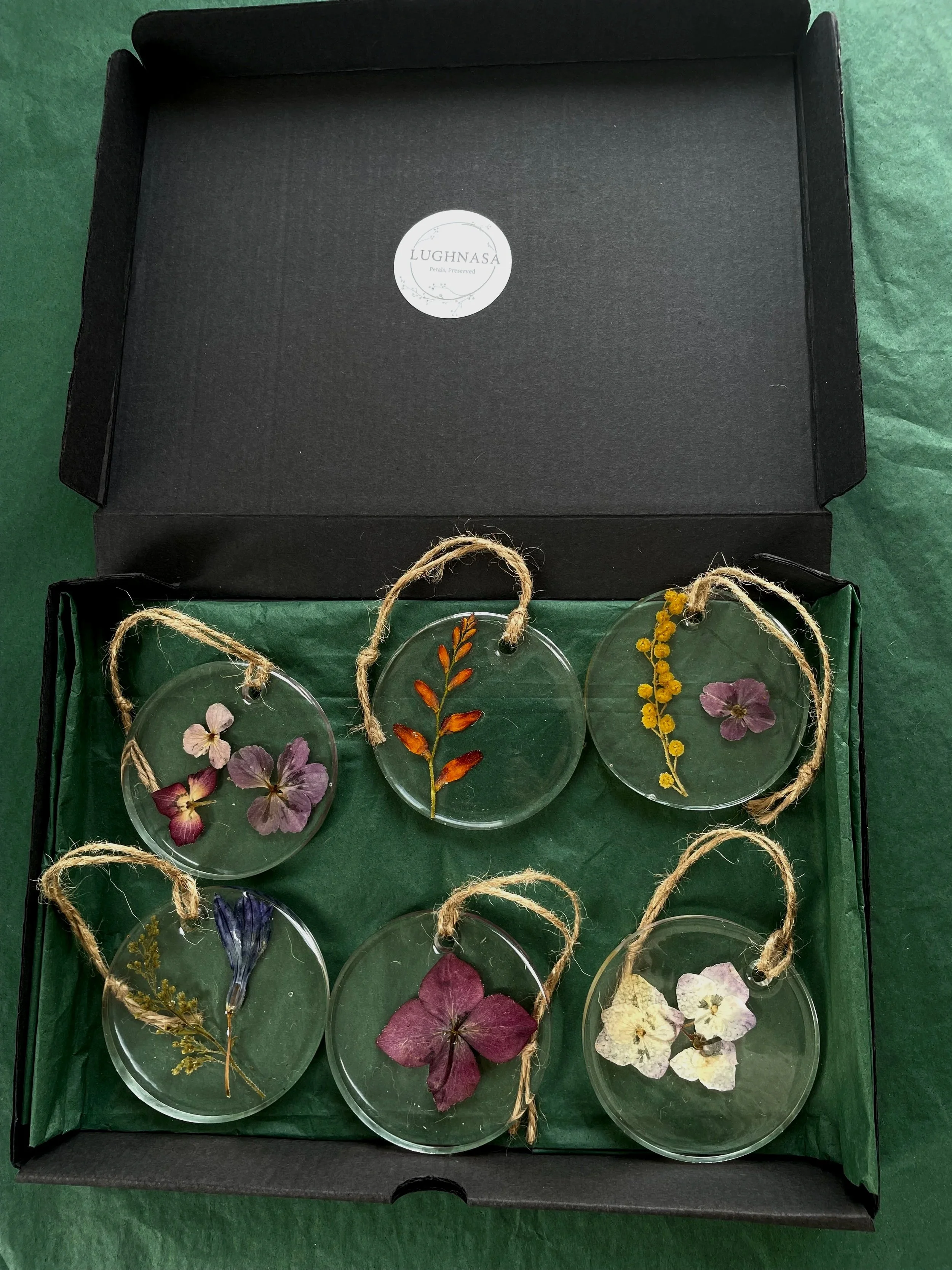 Six glass decorations with pressed flowers inside, arranged in a box with green tissue paper, each with a jute string for hanging.
