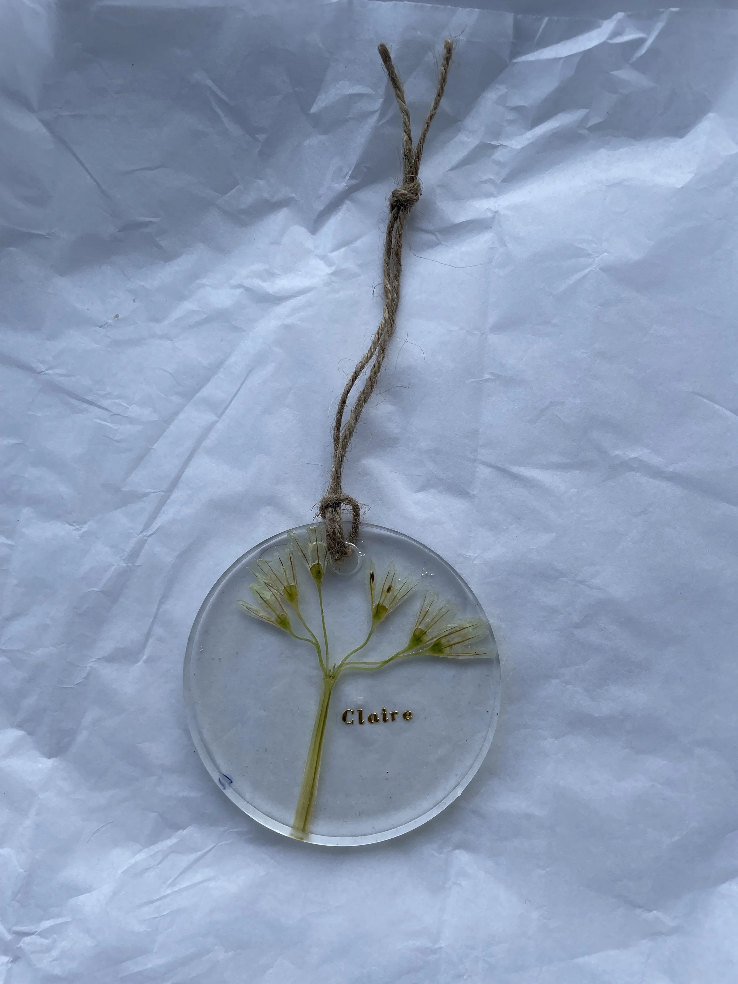 A round glass ornament with a pressed flower inside, featuring a small green stem and multiple tiny yellow flowers. The ornament has the name "Claire" written in gold letters and is attached to a the jute string for hanging, all placed on crinkled wh