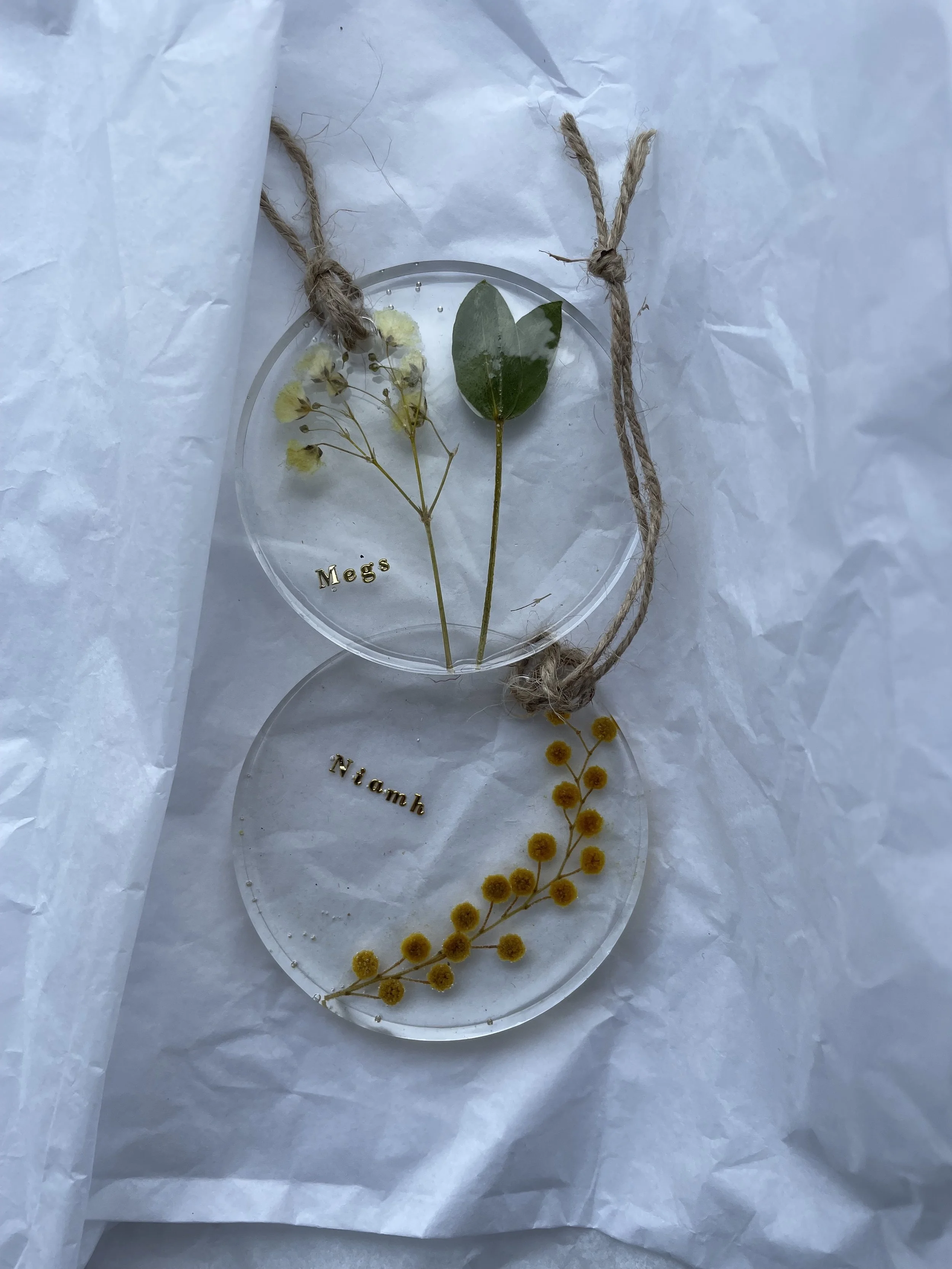 Two transparent circular keepsakes with dried pressed flowers or leaves, tied together with a piece of twine, placed on white tissue paper. Each keepsake has a name: 'Mags' on the top one and 'Niambh' on the bottom one.