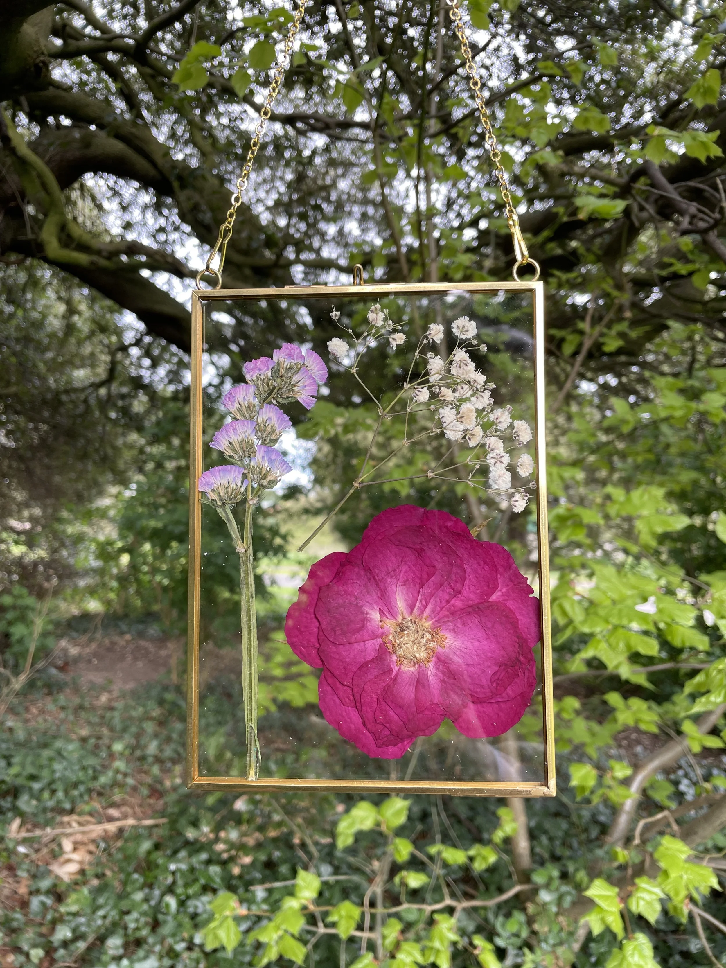 A hanging frame with pressed purple, white, and pink flowers outdoors among green trees and leaves.