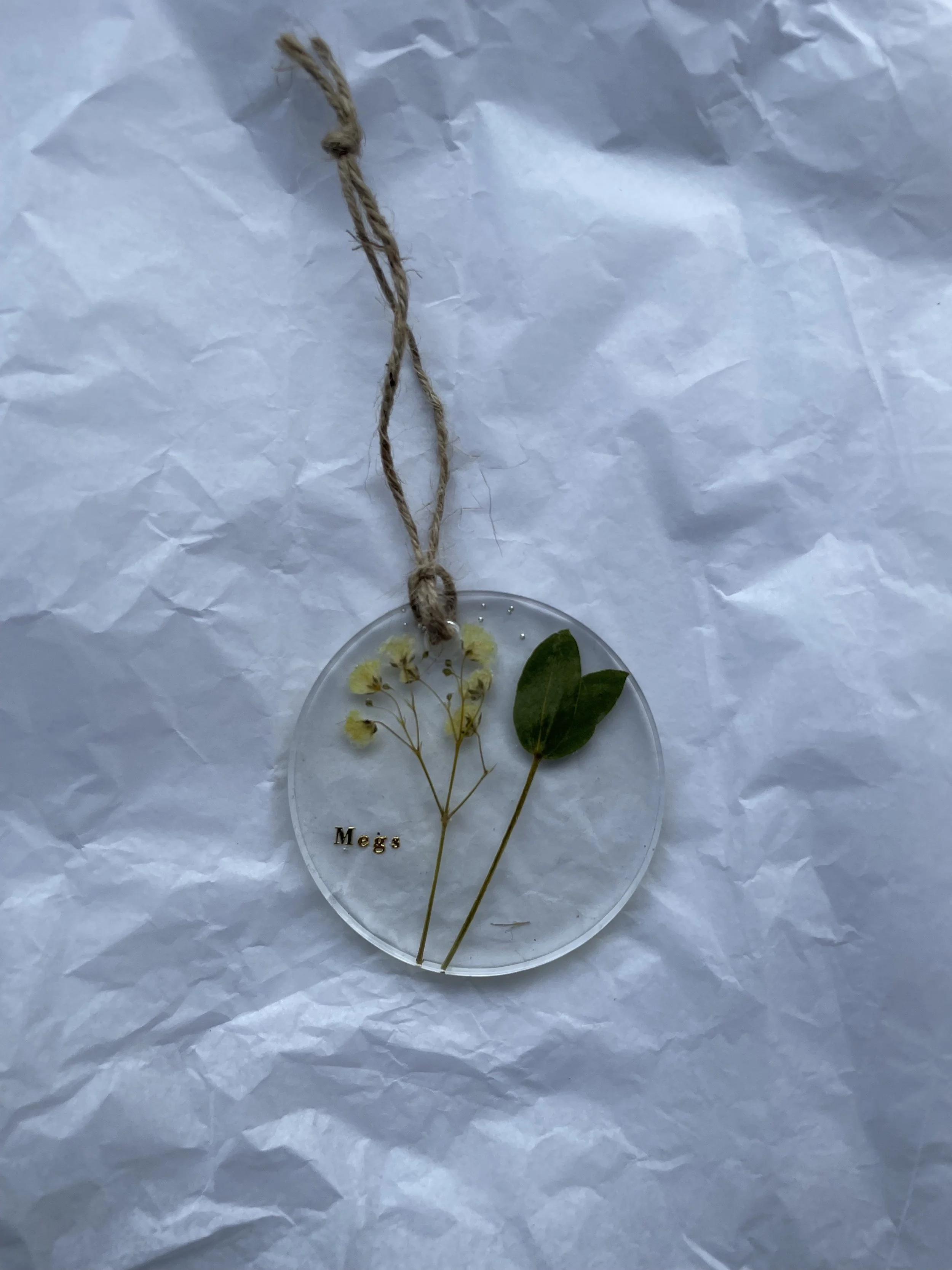 A round clear ornament with yellow flowers and green leaves inside, attached to a tan string, resting on crinkled white paper, with the name 'Megs' in small black letters inside.