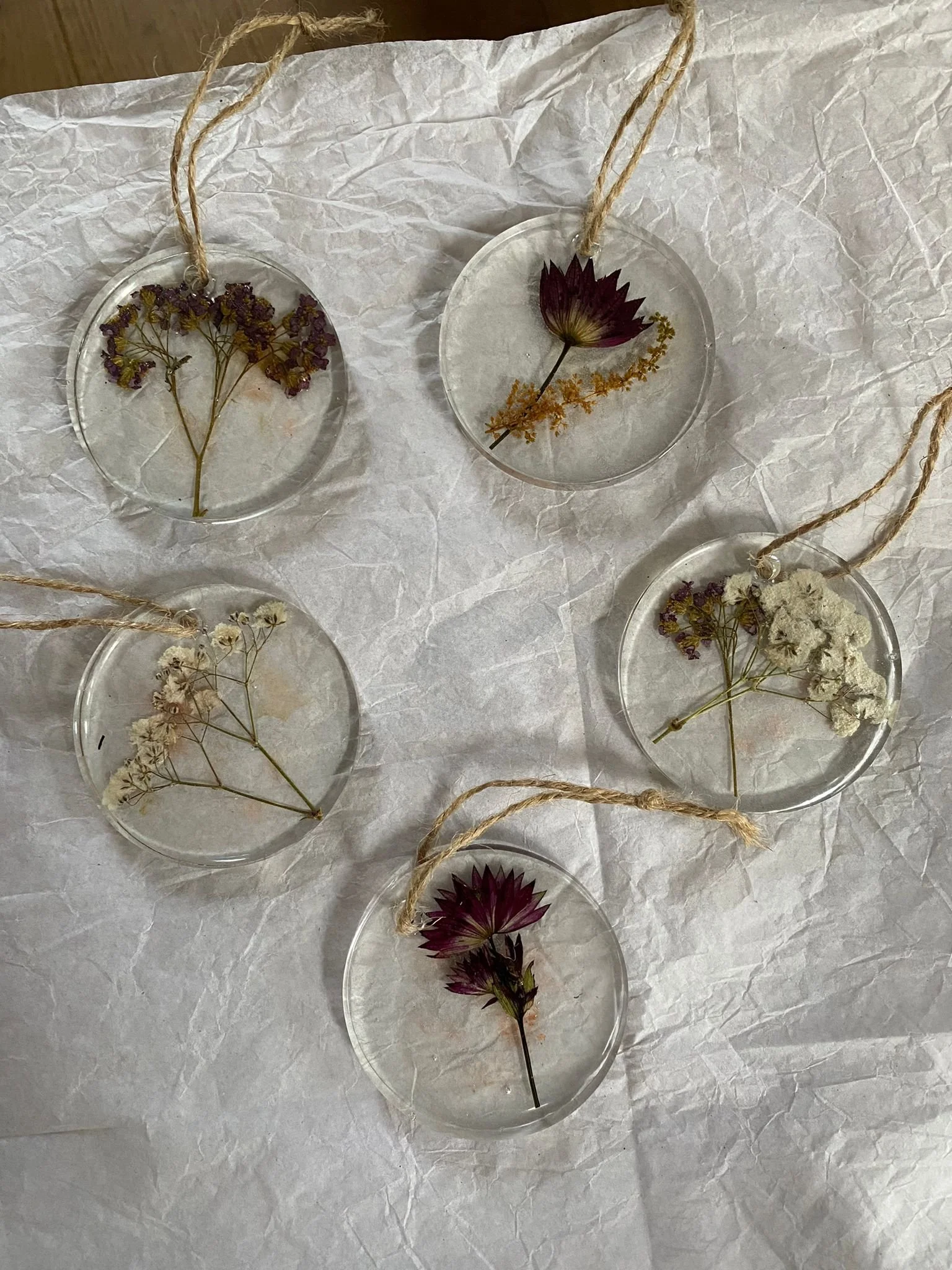 Six circular resin ornaments with dried pressed flowers inside, attached to jute strings. The flowers include purple, yellow, white, and dark pink blooms, arranged in various designs. They are placed on crinkled white paper.